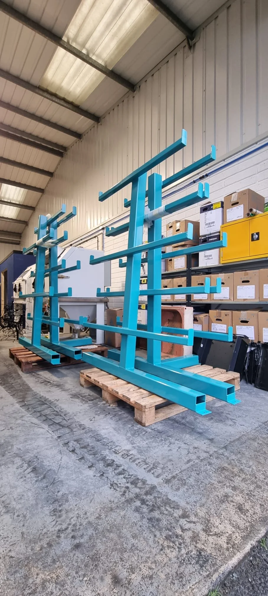 Two large, blue metal powdercoated shelving units on wooden pallets in a storage area with cardboard boxes and other equipment in the background.