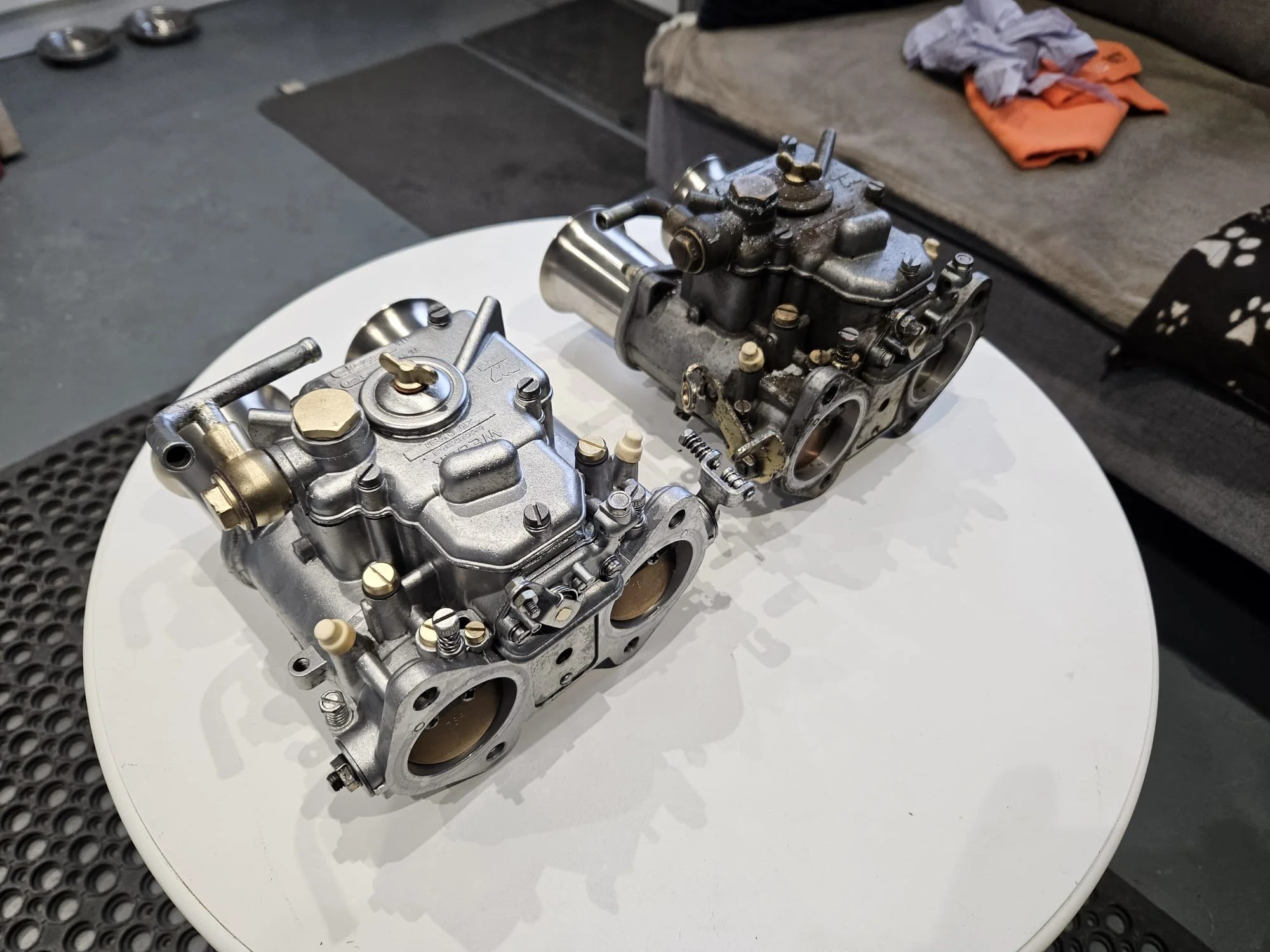 Two metal carburetor components placed on a white circular table. one old and one fresh.