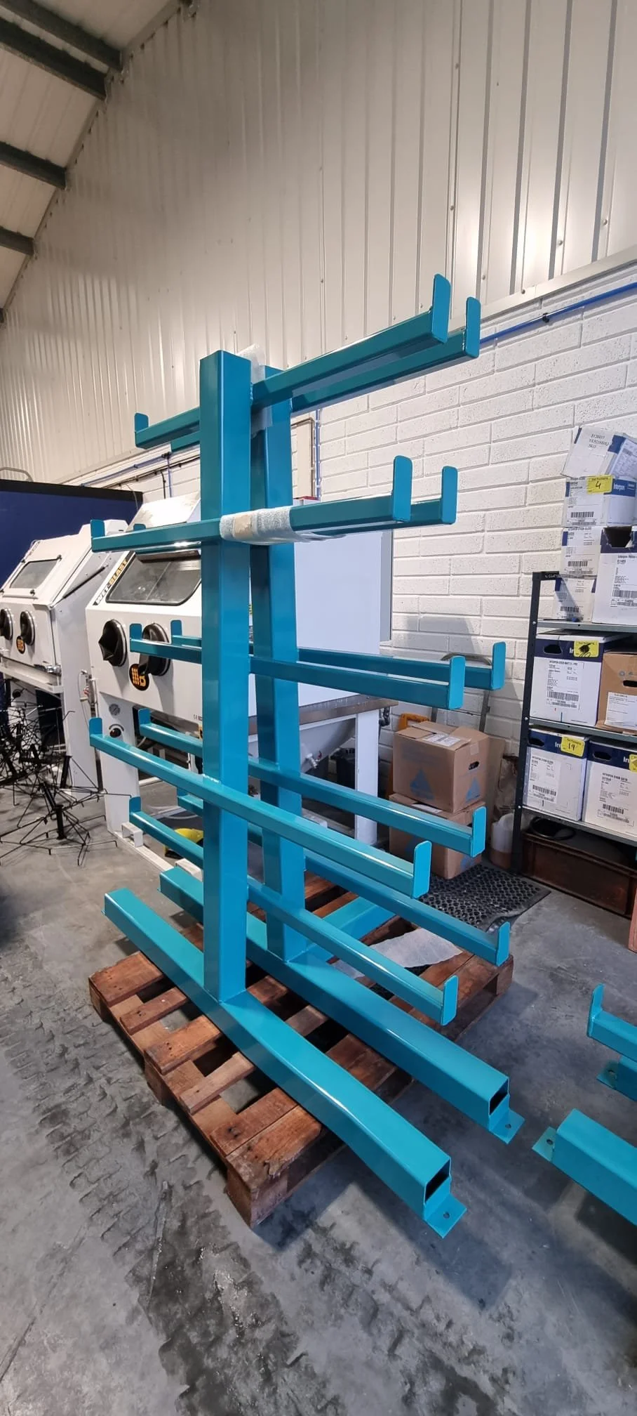 A blue powdered metal storage rack with five shelves, placed on a wooden pallet in an industrial warehouse with white brick walls and carting machines in the background.