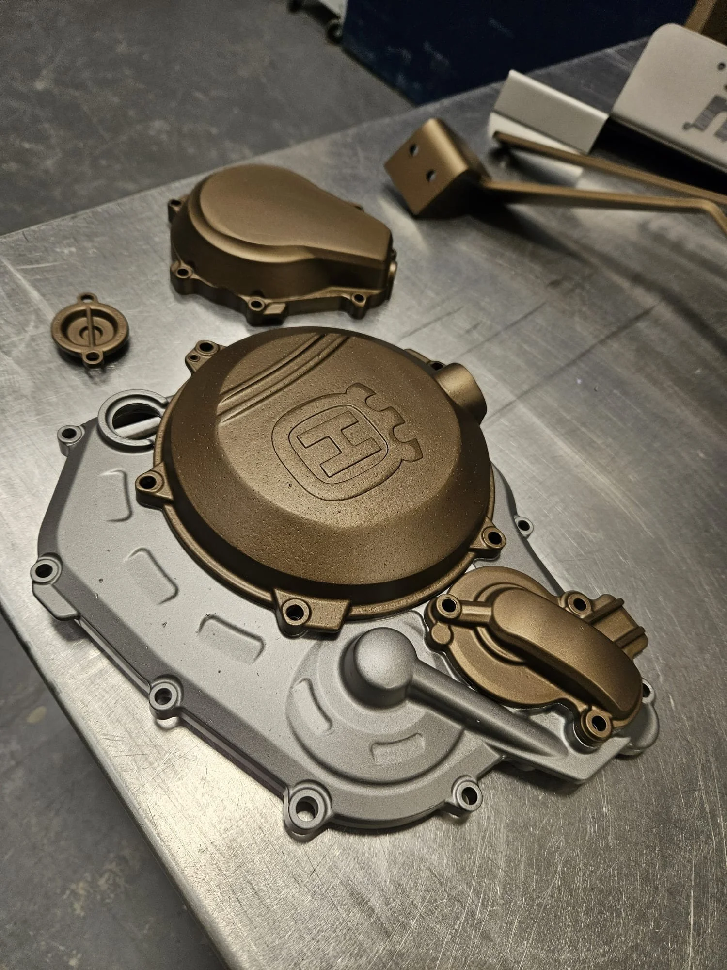 Close-up view of a disassembled motorcycle engine part, including a silver engine casing, a gold-colored cover with a logo, and other smaller components, placed on a metallic work surface.