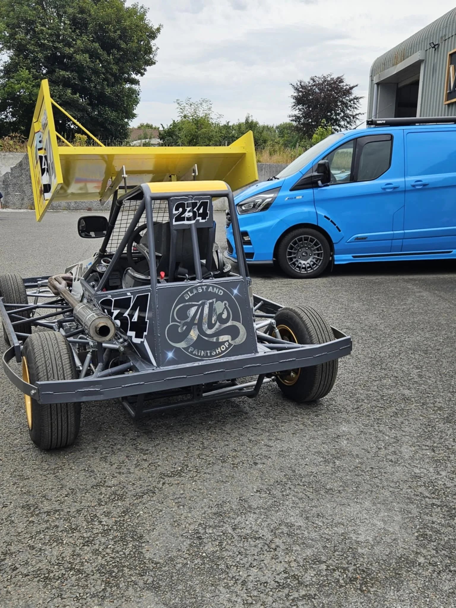 A go-kart with number 34 and a large black and gray logo reading 'Al's Blast and Paintshop' on the front, parked on a gravel surface next to a blue cargo van with a yellow rear wing attached.