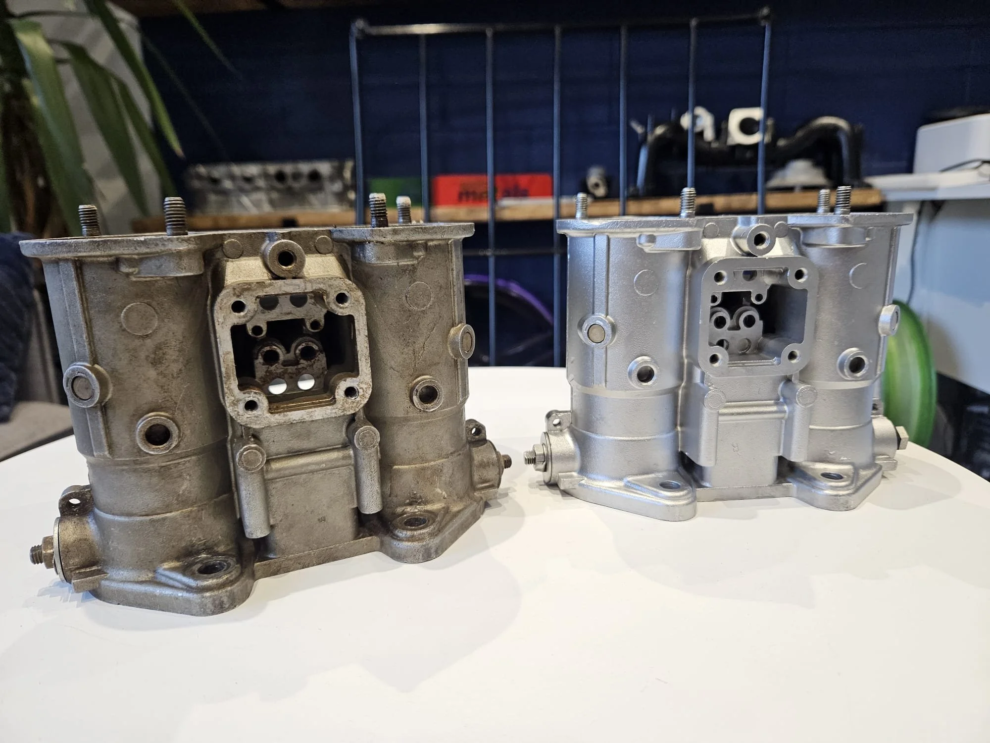 Two motorcycle carburetors, one old and dirty on the left, and one new and clean on the right, placed on a white surface before and after of vapor blasting and cleaning work.
