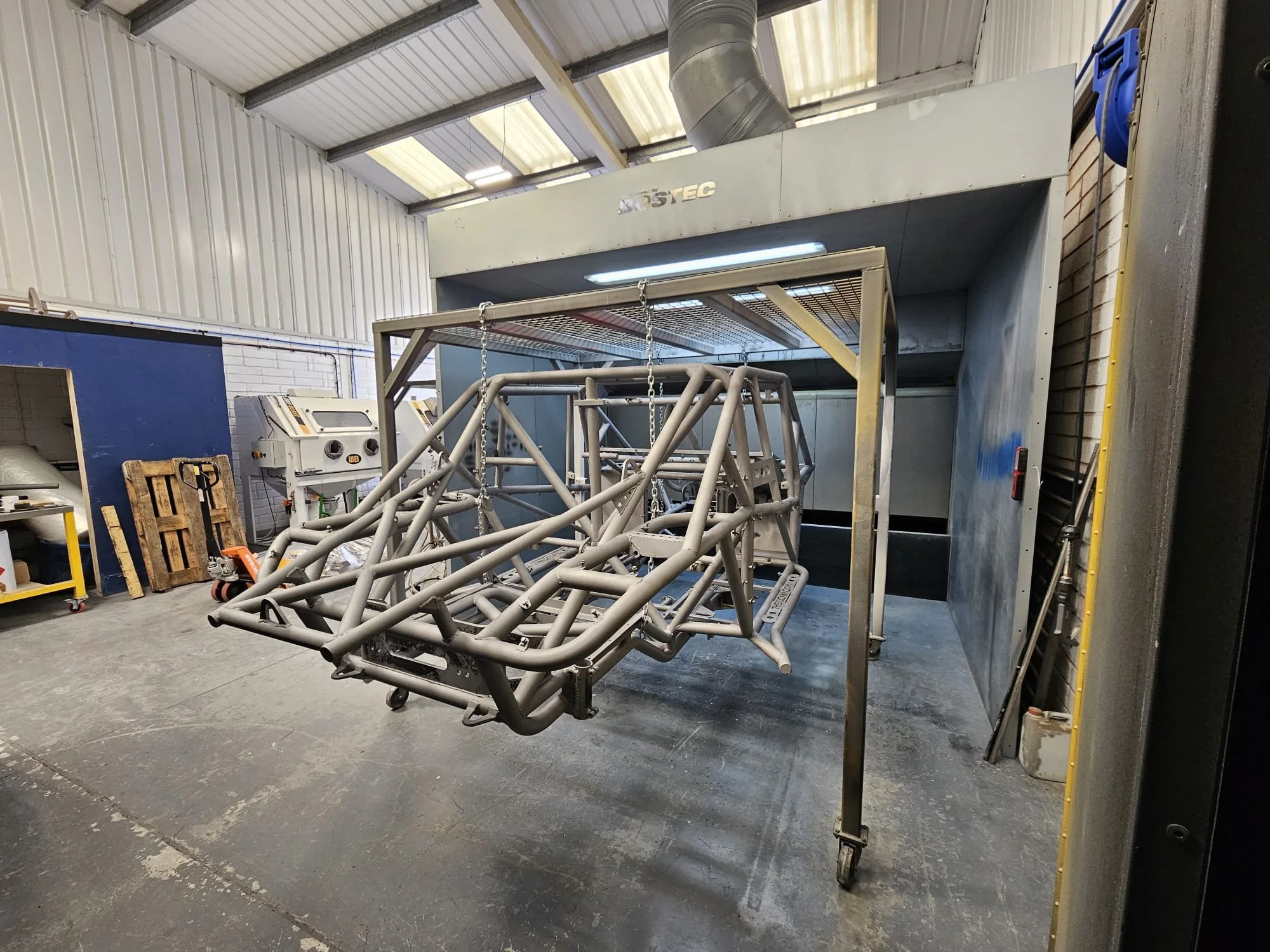 A metal racing car frame suspended in a paint booth with a ventilation hood above, inside a workshop with white and blue walls, pallets, and equipment.