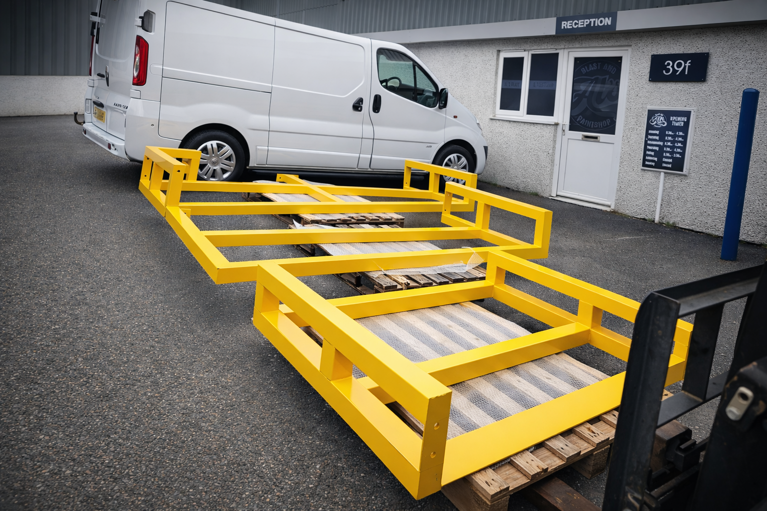 A white van parked in front of a building with a reception area. A yellow powder coated metal safety barrier surrounds a wooden pallet on the ground, which appears to be used for loading or unloading.