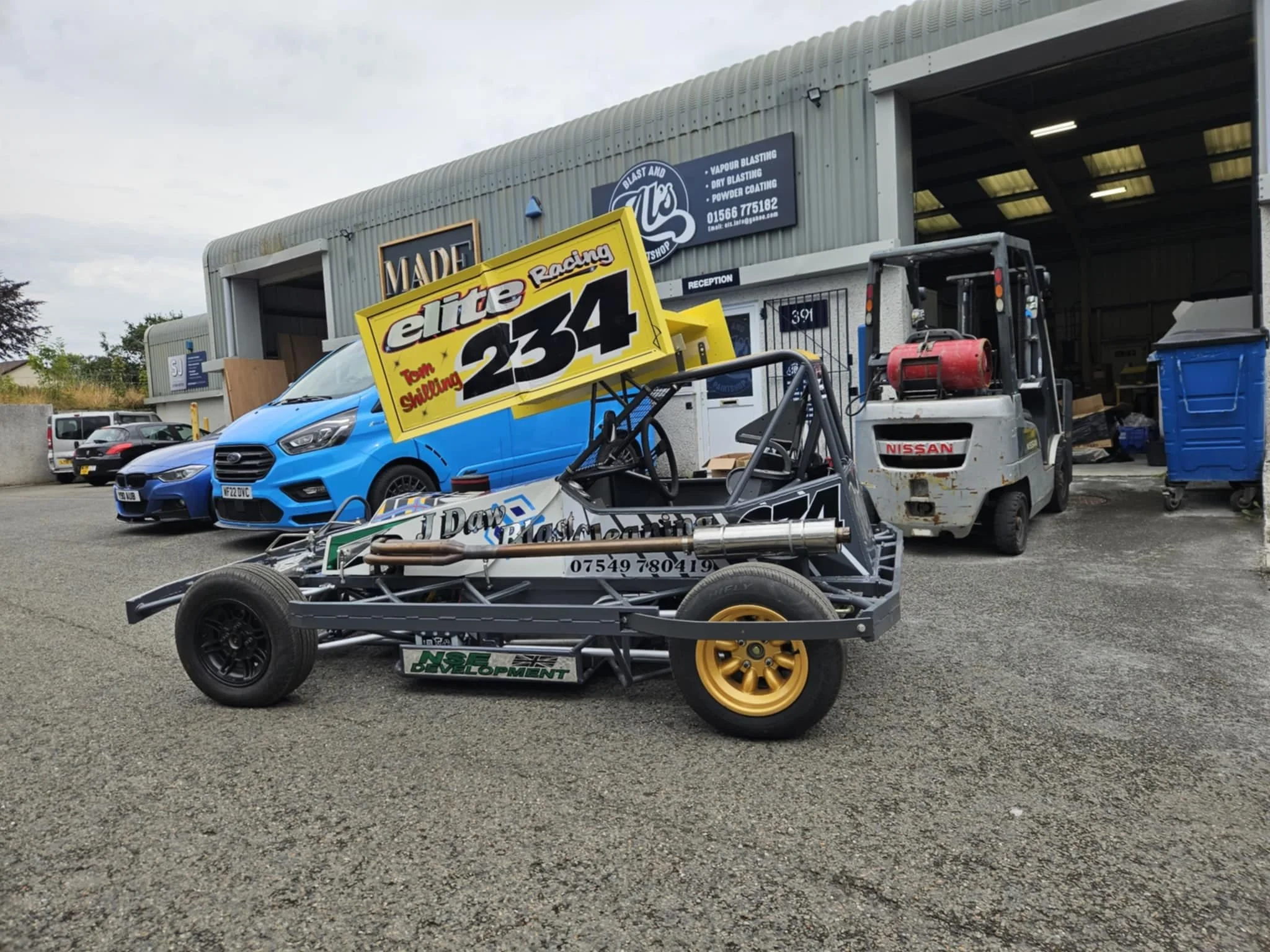 A go-kart parked outside a workshop, with a blue car and a forklift in the background.