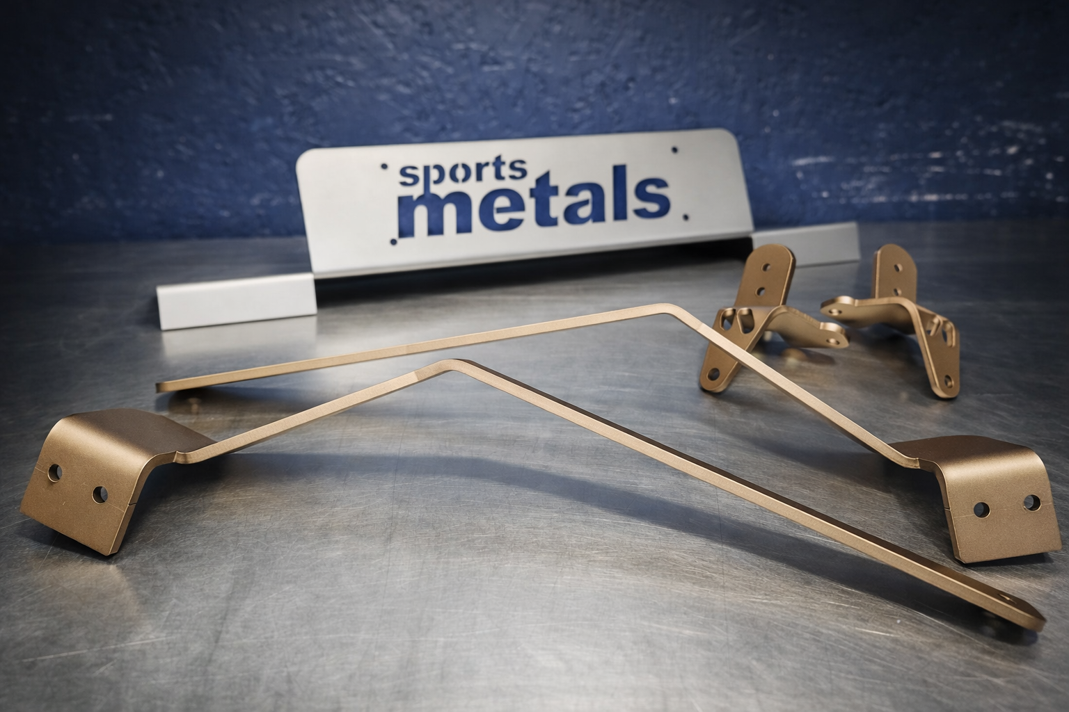 Gold-colored metal sports car roll cage parts, with a blue and white sign reading 'sports metals' in the background on a gray surface.