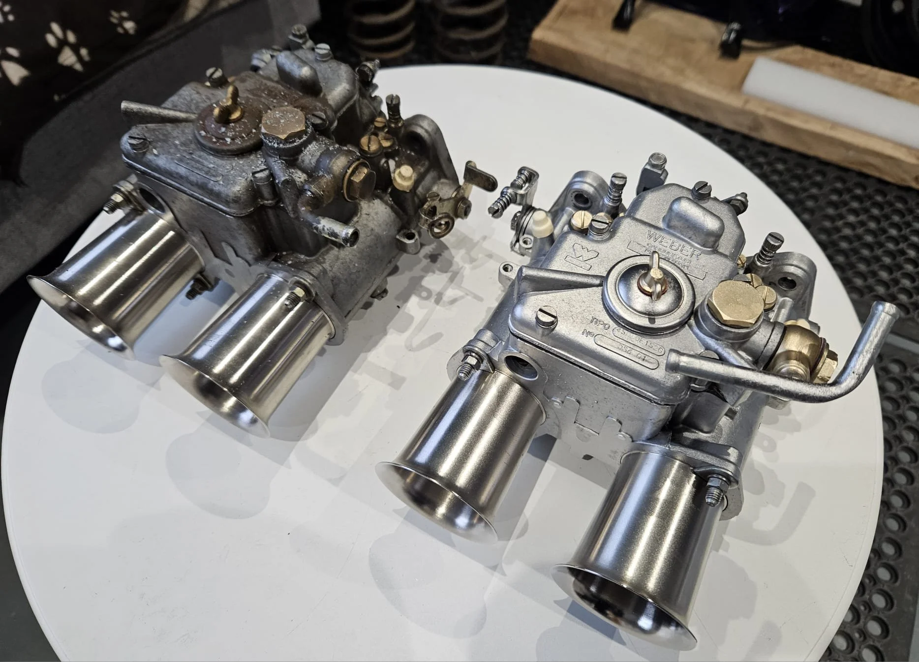 Two mechanical carburetors with aluminum bodies and metal nozzles on a white circular surface showing a before and after vapour blasting process. 
