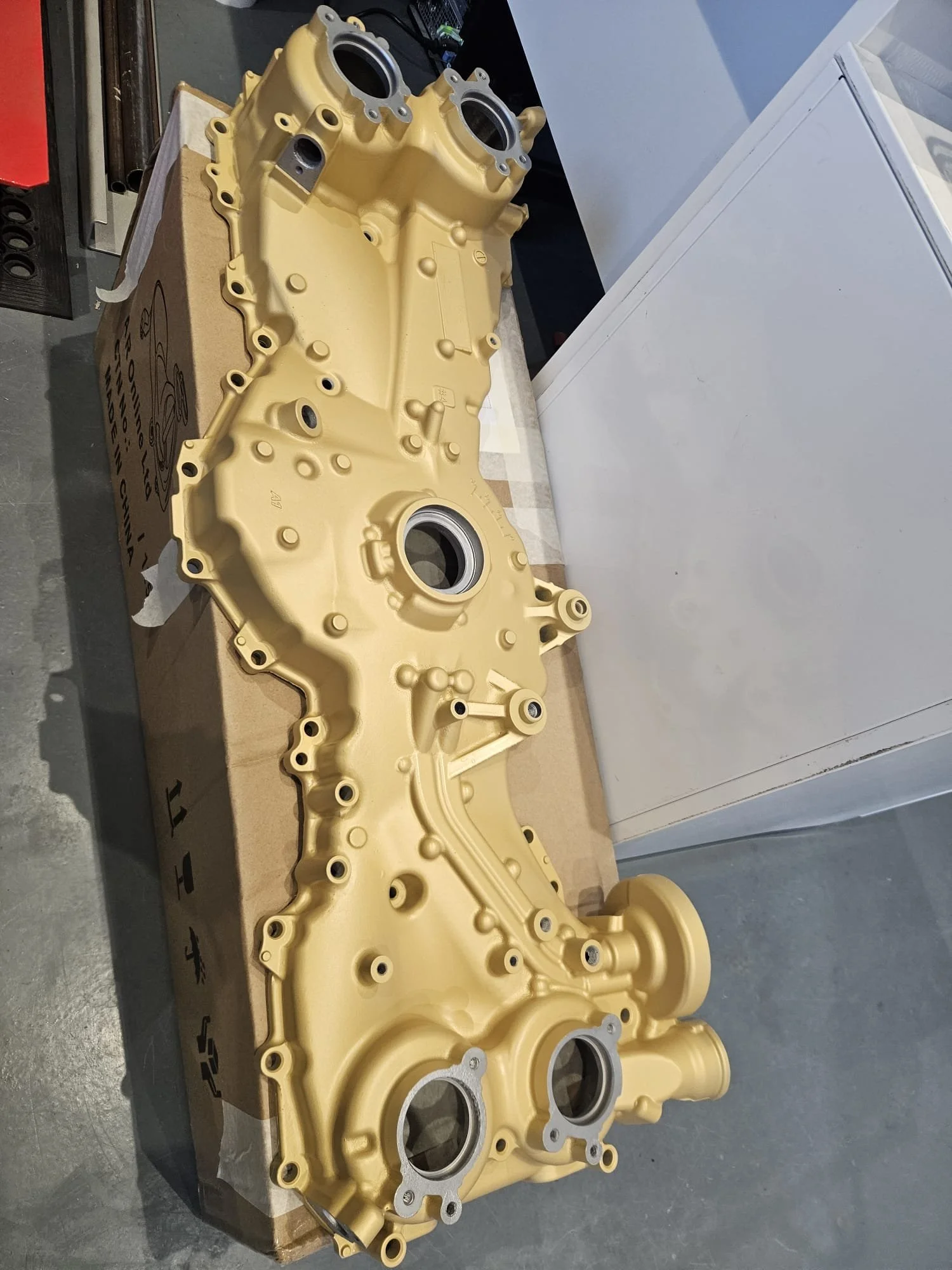 A yellow automotive engine part, likely a valve cover, is resting on a cardboard box on a workbench.