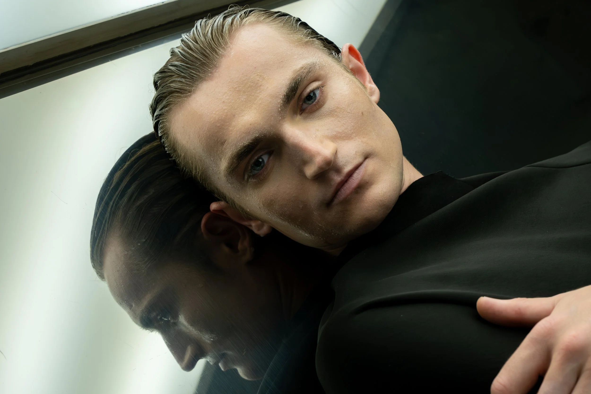 Close-up of a young man with blonde hair and blue eyes lying on a metallic surface, gazing into the camera.