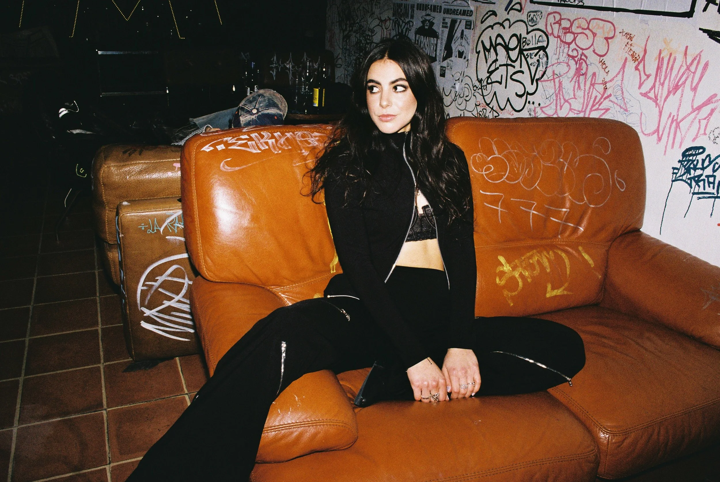 A young woman with long dark hair, wearing a black outfit with zippers, sitting cross-legged on a brown leather couch in a graffiti-covered room.