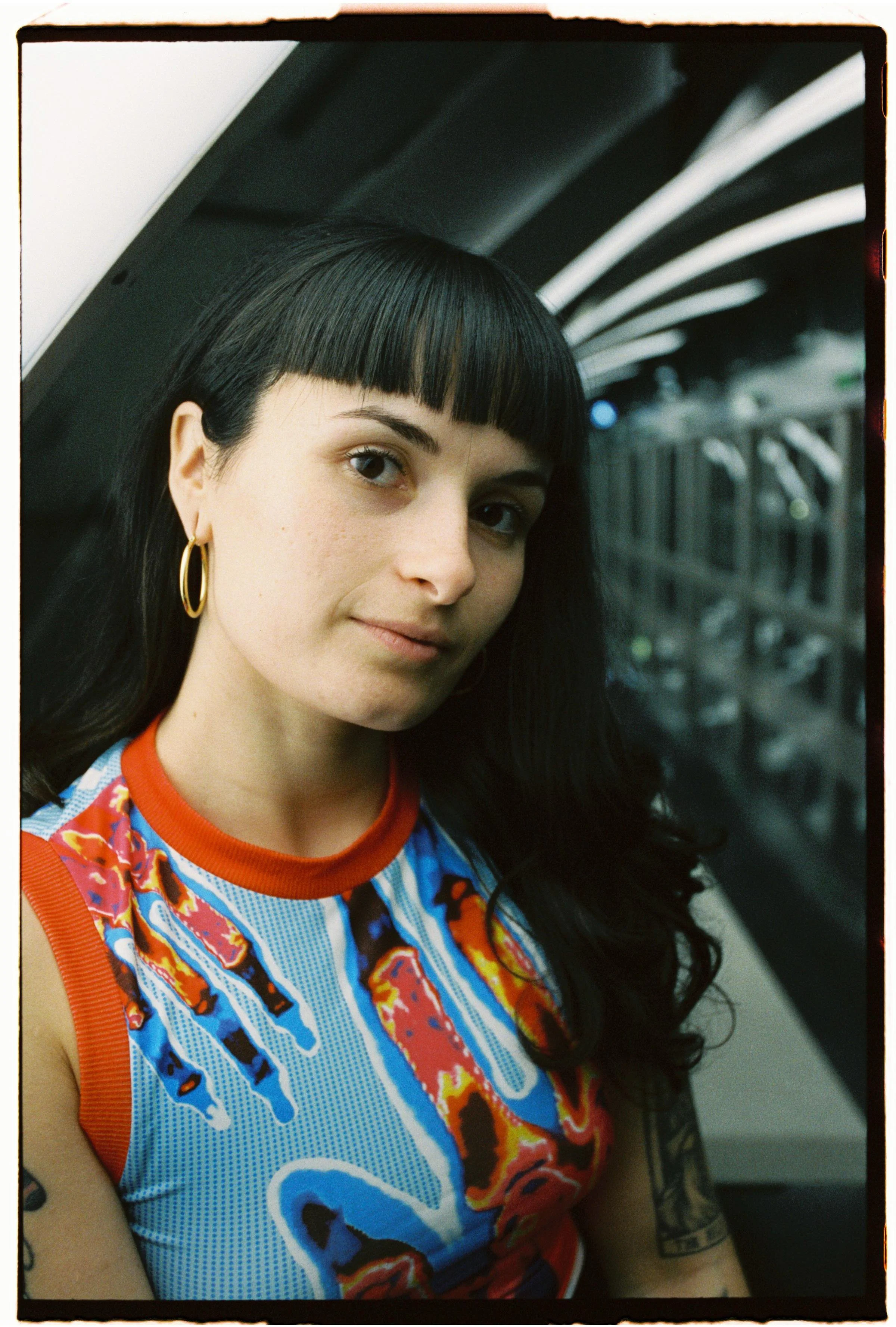 A woman with black hair, cut in a blunt bob with bangs, wearing gold hoop earrings and a colorful tank top, sitting in a subway train, looking at the camera.