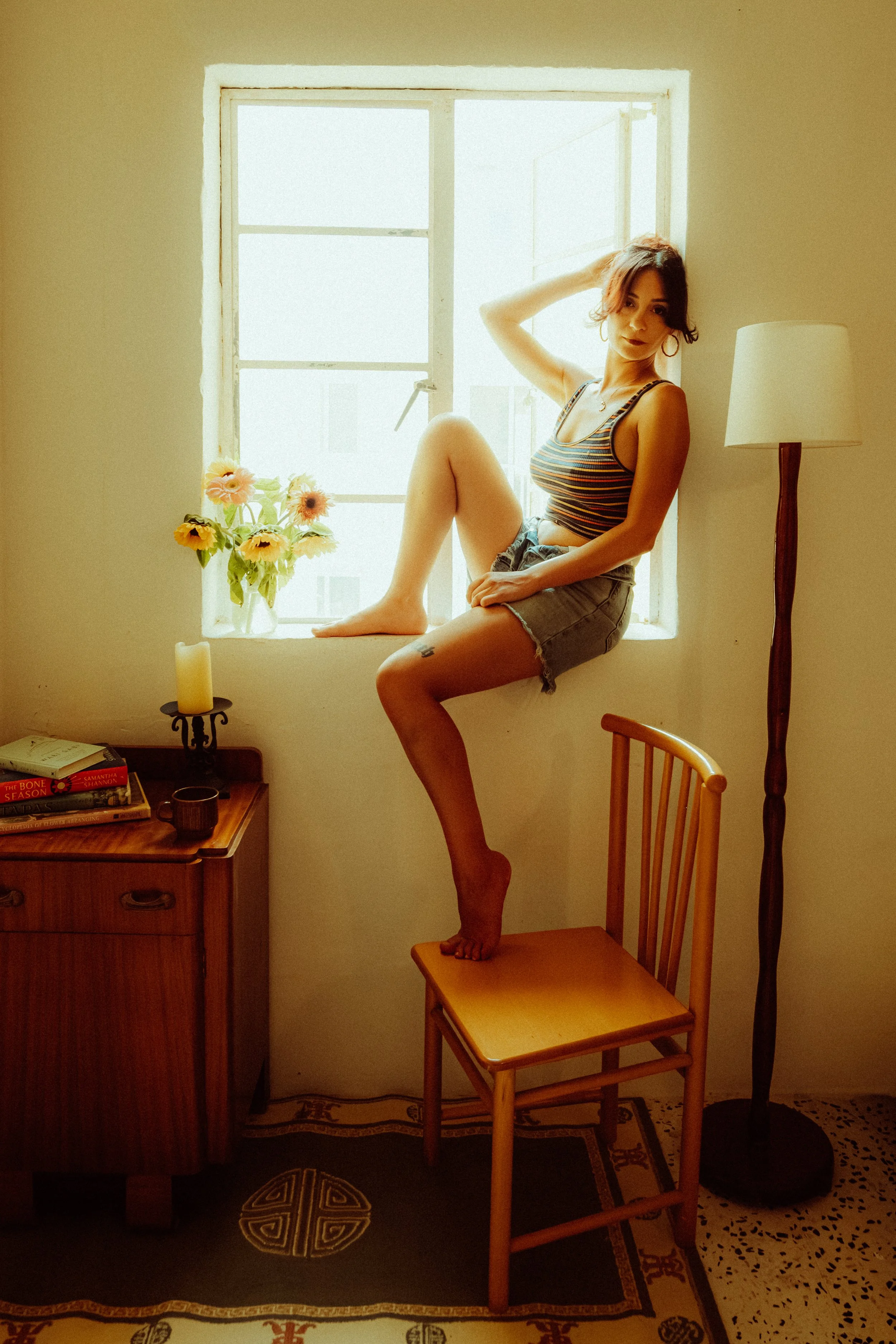A woman sitting on a window ledge near a window with open shutters, with a lamp beside her, flowers in a vase, and a small table with books below.