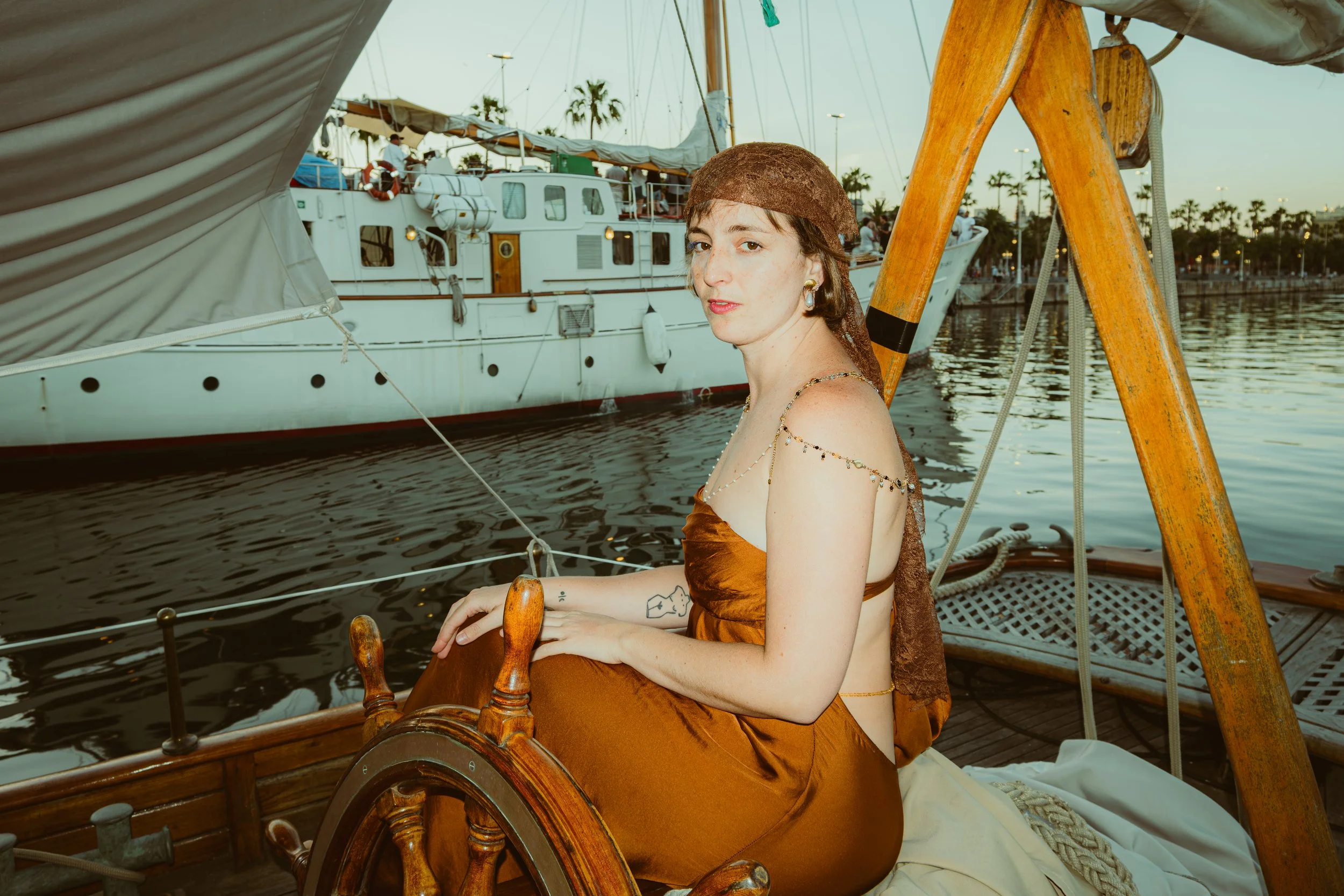 A woman with tattoos wearing a brown dress and headscarf sitting on a wooden boat, with a white yacht, palm trees, and other boats in the background at sunset.
