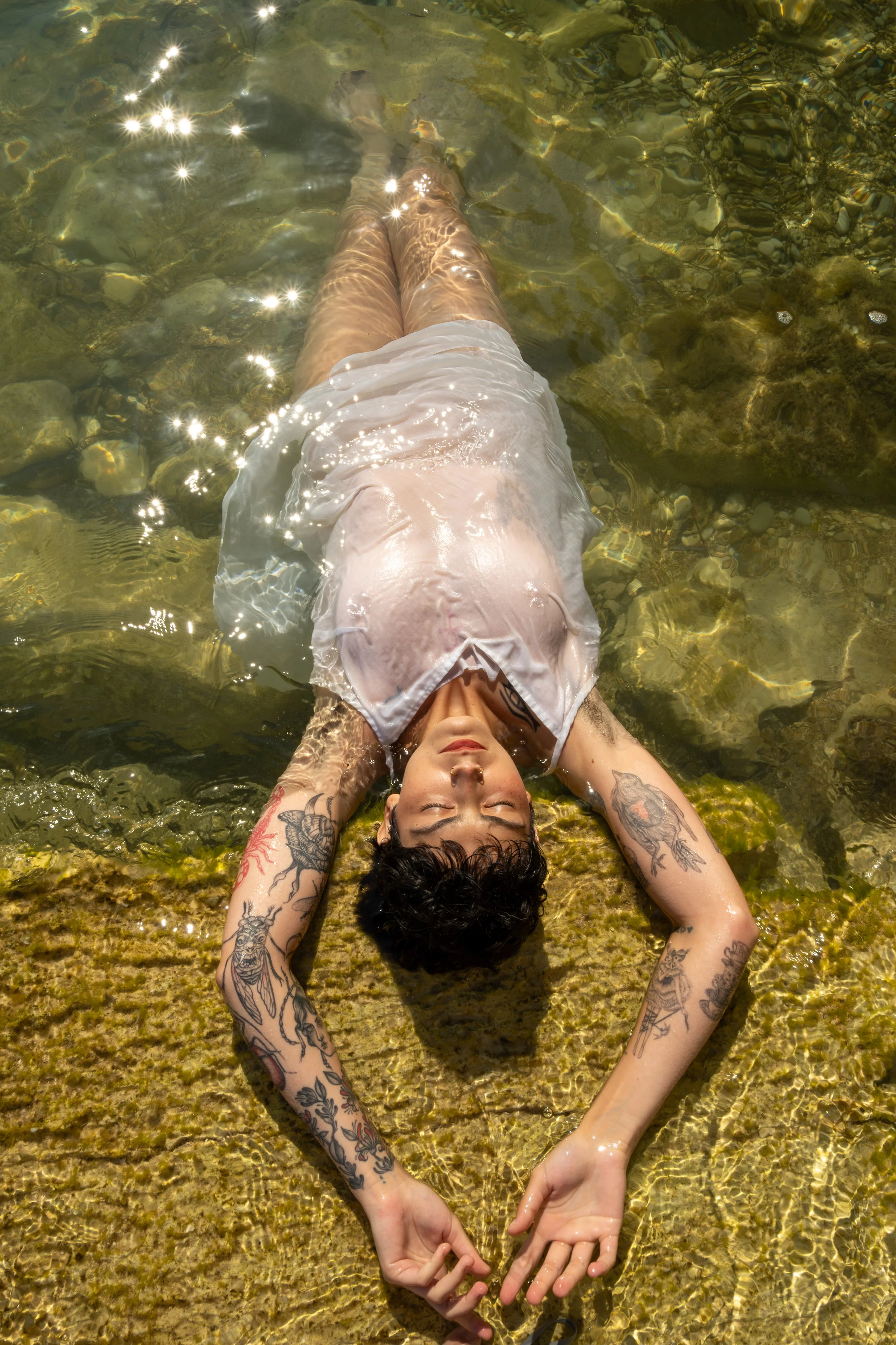 A person with tattoos is lying on their back in shallow, clear water on a sunny day, with arms stretched above their head and eyes closed.