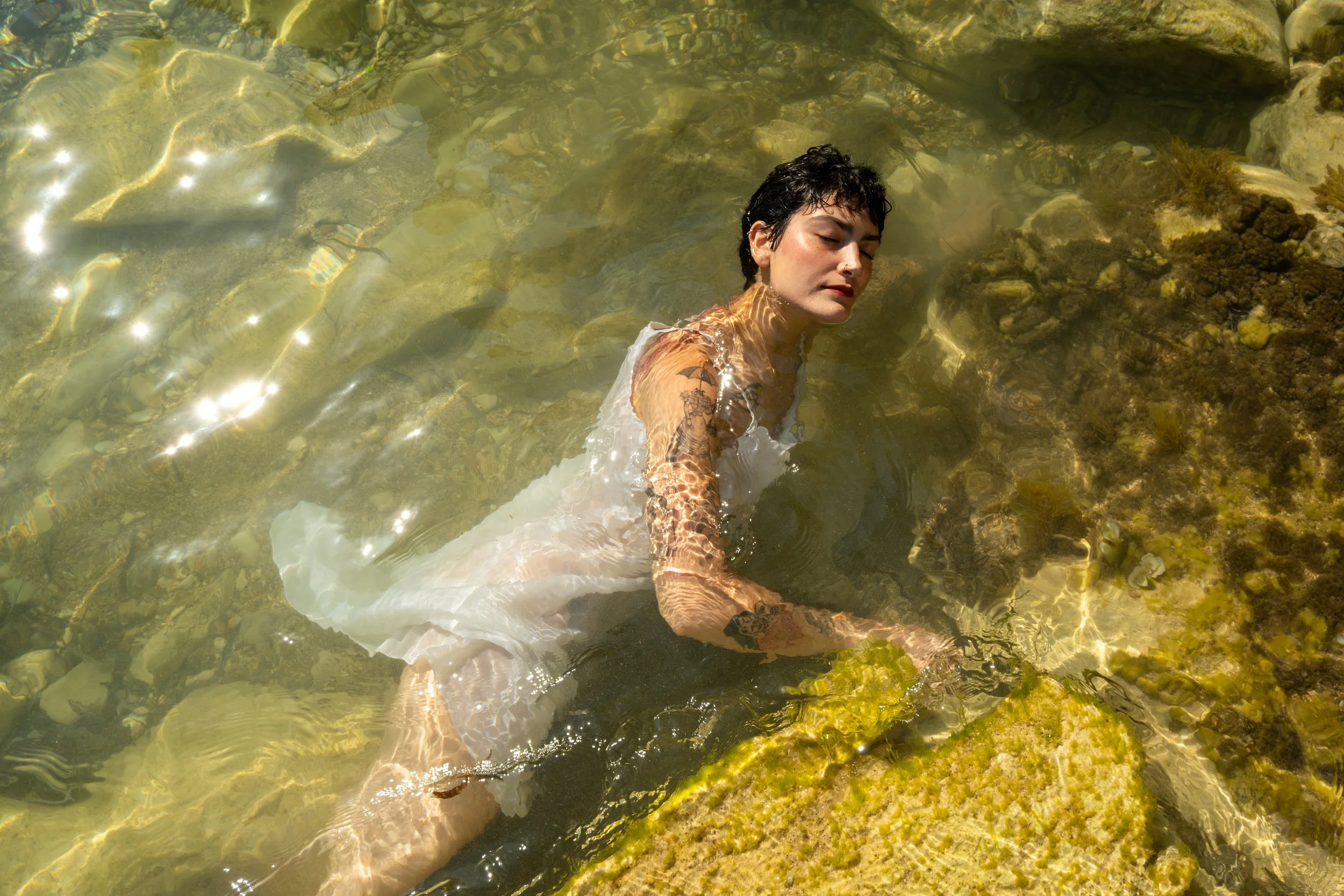 A young woman with short dark hair and visible tattoos relaxing in clear, shallow water among rocks and algae, with sunlight reflecting on the surface.