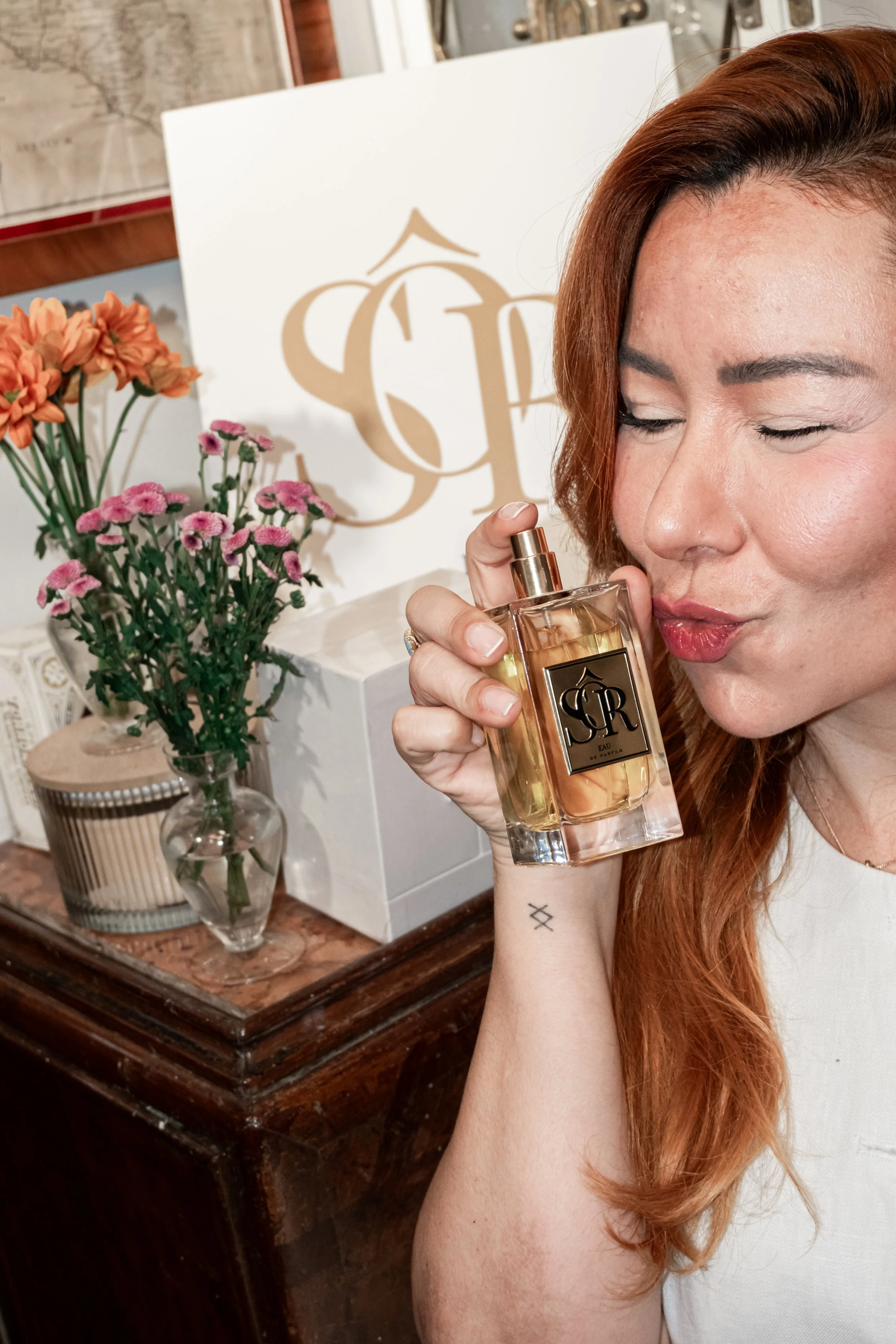 A woman with red hair holding a perfume bottle, closing her eyes and smiling softly. In the background, there is a floral arrangement with orange and pink flowers and a white box with a monogrammed logo.