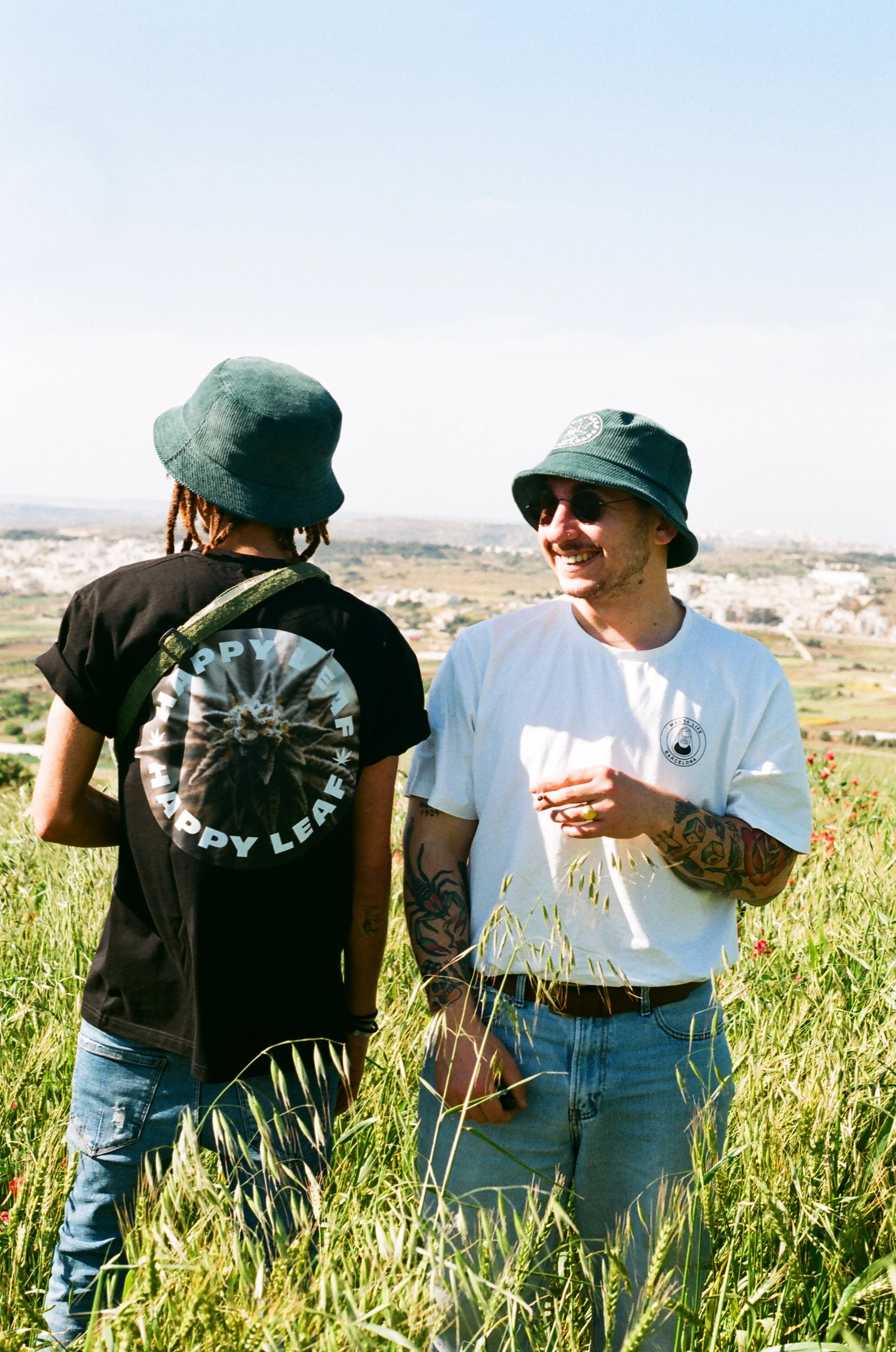 Two young men standing in a grassy field, smiling and talking, with a scenic landscape and city in the background. Both wear bucket hats, one with sunglasses, casual clothing, and tattoos visible on their arms.