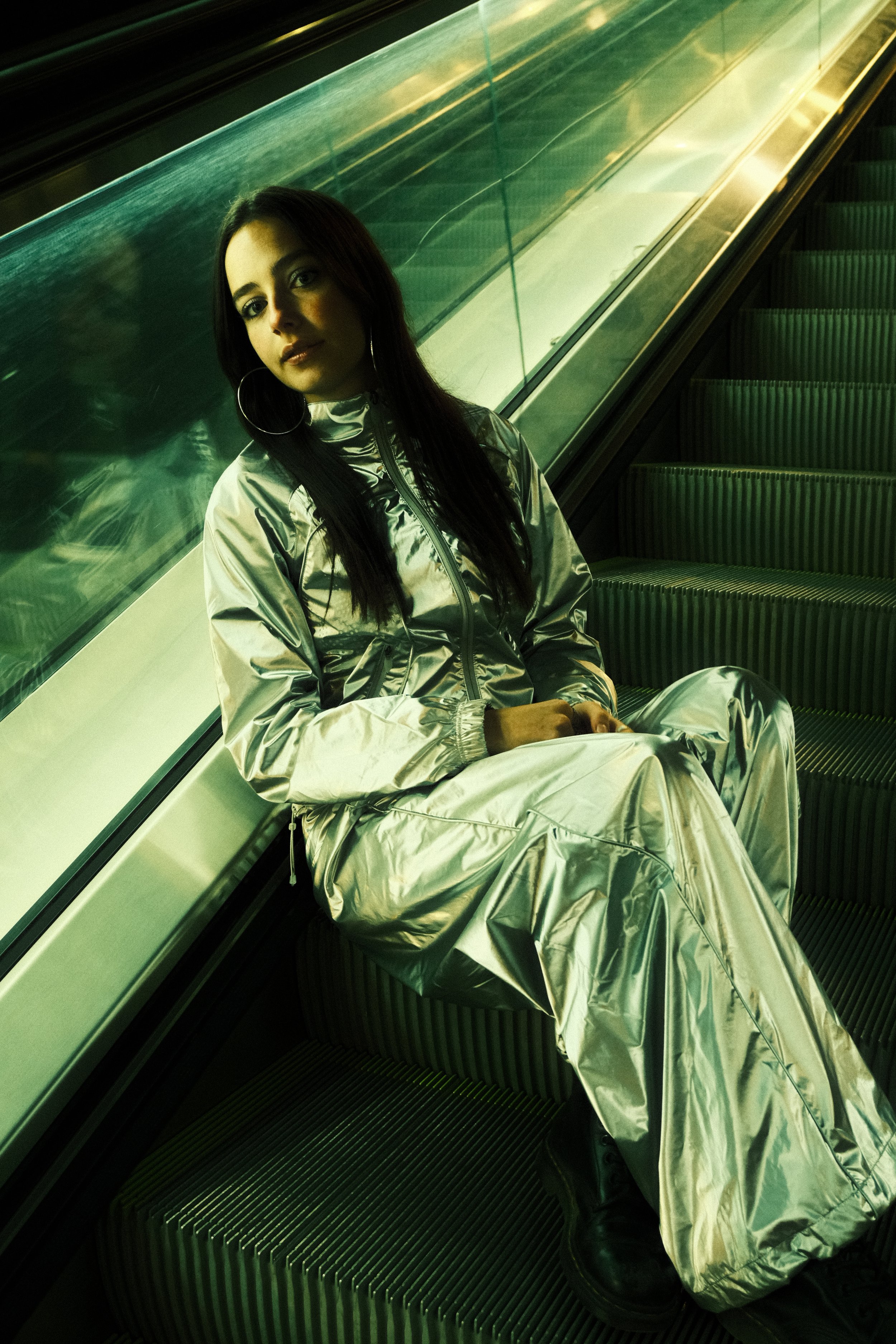 A woman with long dark hair, wearing a shiny silver tracksuit and large hoop earrings, sitting on an escalator with green lighting, next to a glass railing.