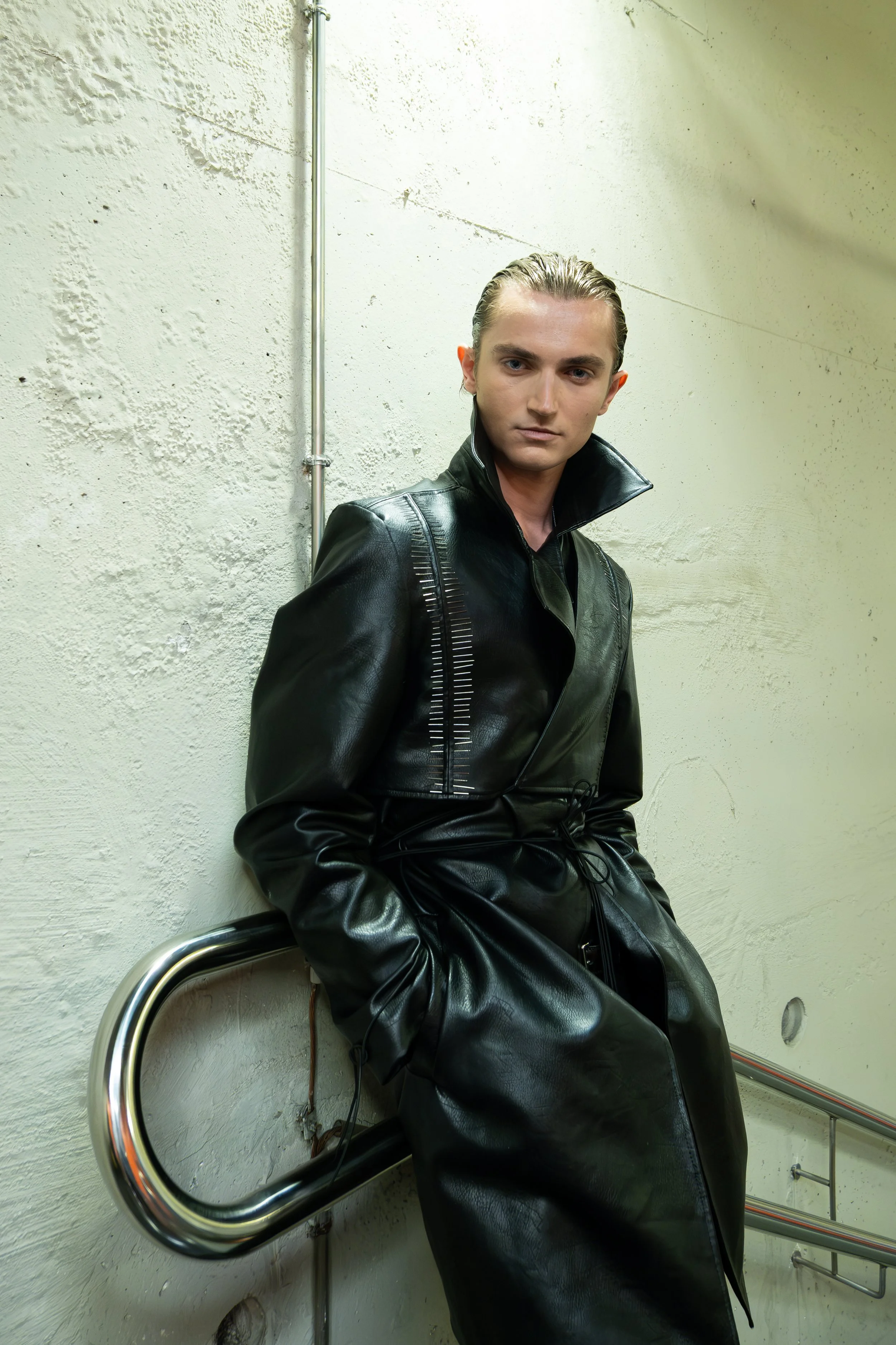 A young man with slicked-back hair and a serious expression, wearing a black leather jacket, sitting against a rough white wall with metal pipes and railing nearby.