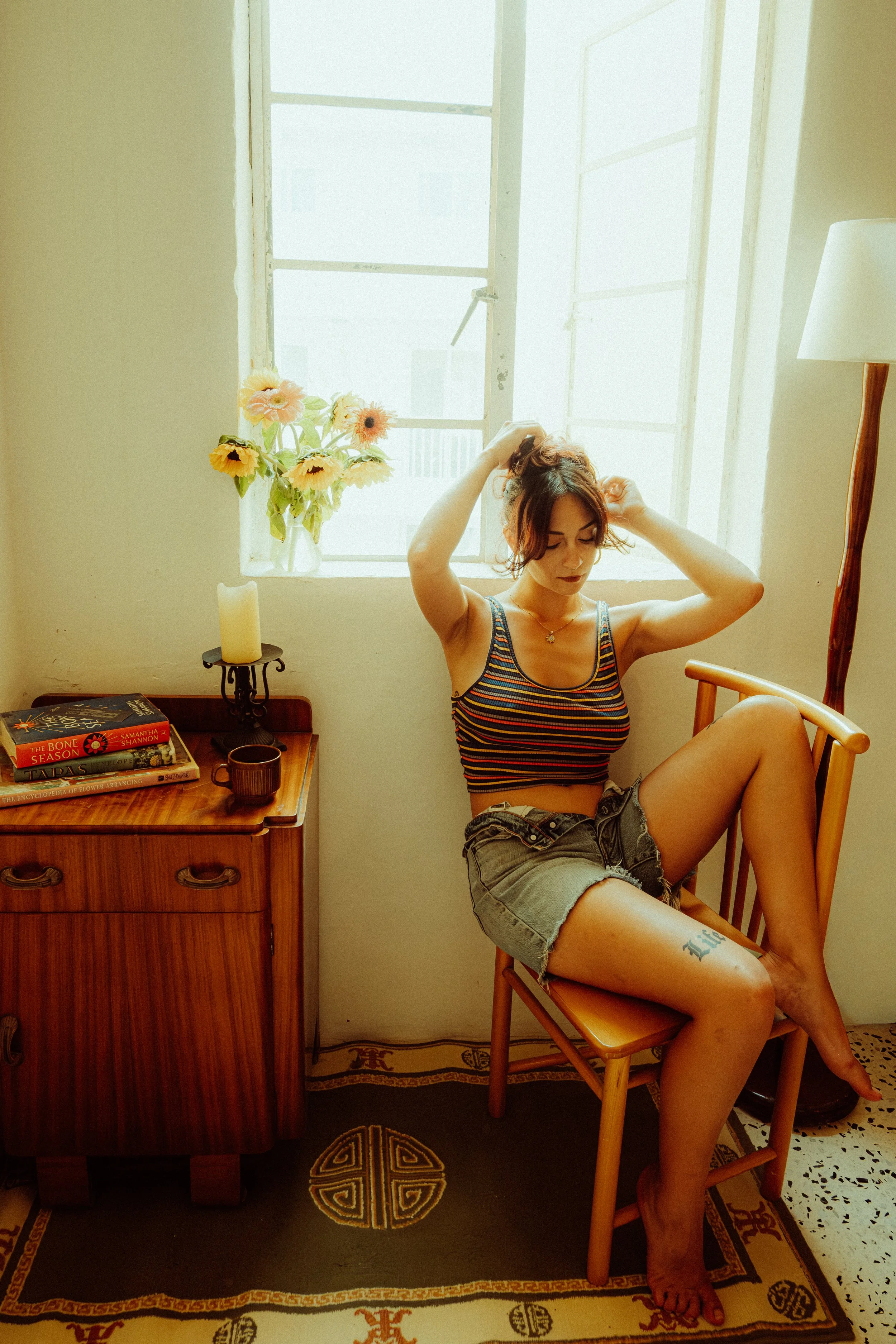 A young woman with dark hair, wearing a striped tank top and denim shorts, sitting on a wooden chair. She is adjusting her hair and looking down. There is a window behind her with sunlight streaming in, a vase of flowers, a candle, and a couple of bo