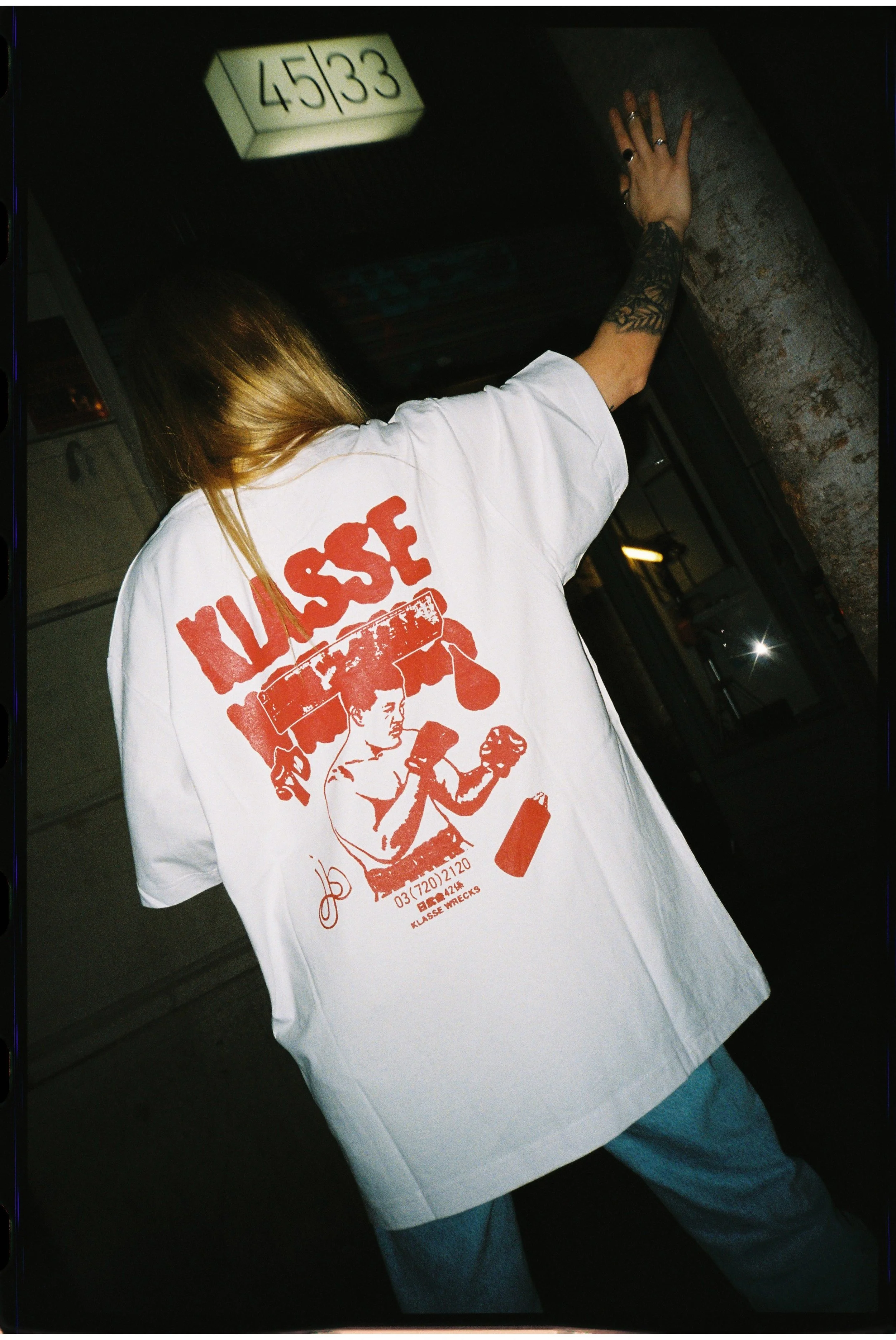 Person wearing a white t-shirt with red graphic print on the back, raising one arm, standing next to a brick column in a dark indoor setting.