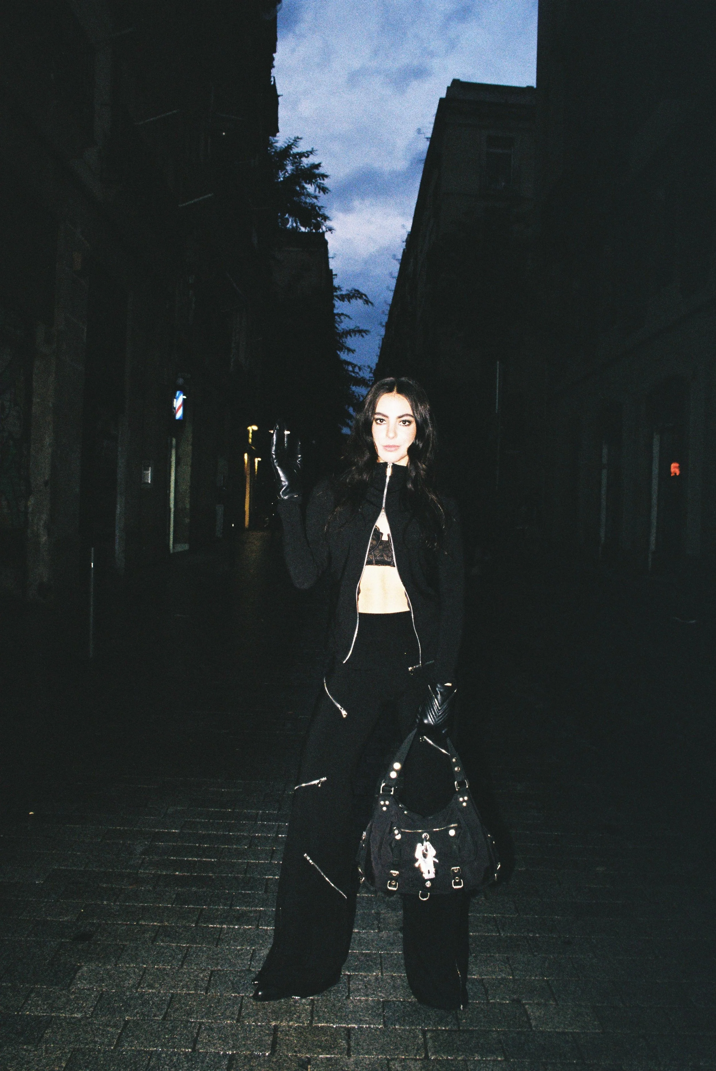 A woman dressed in black stands on a dark city street during dusk, holding a black handbag and wearing gloves, with buildings and a cloudy sky in the background.