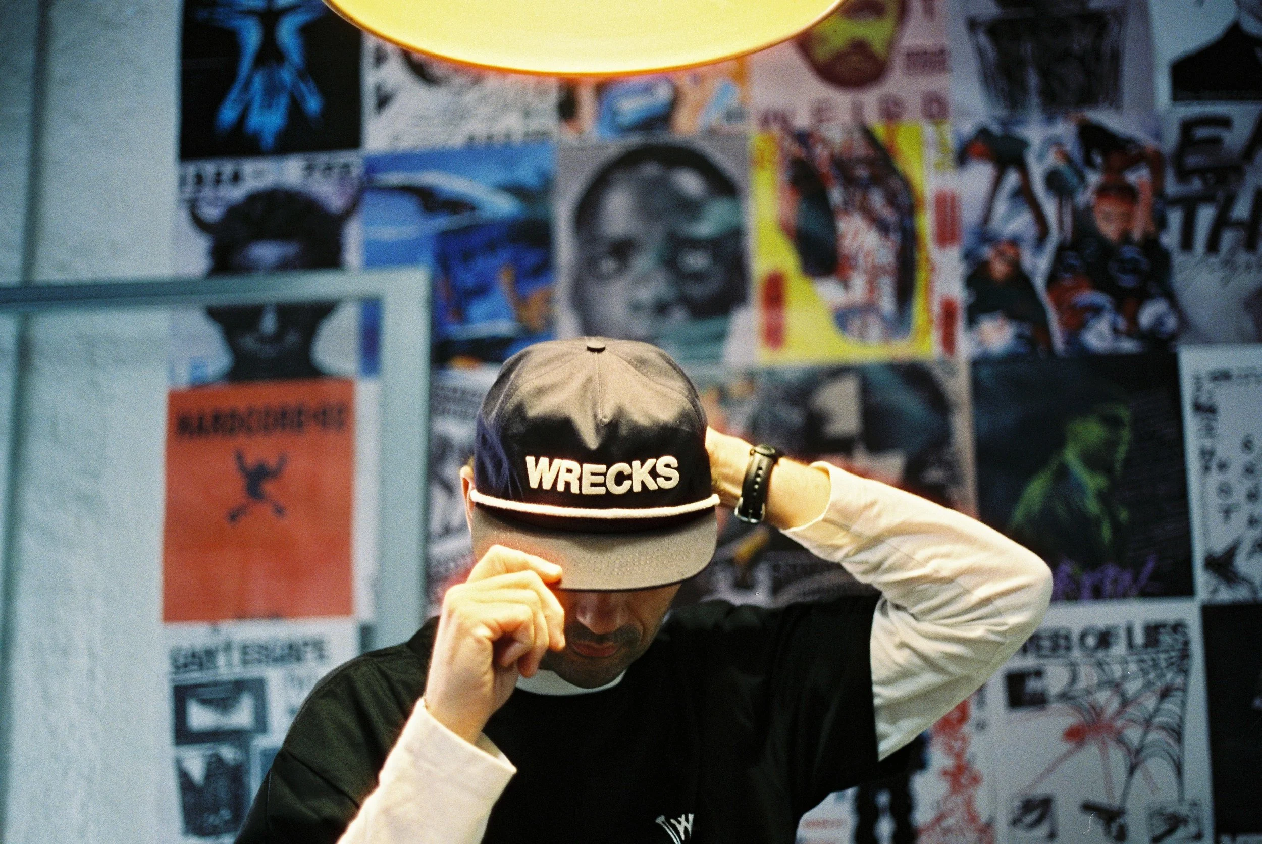 Person wearing a black hat with 'WRECKS' embroidered on the front, adjusting the hat in a room with collage-style posters on the wall.