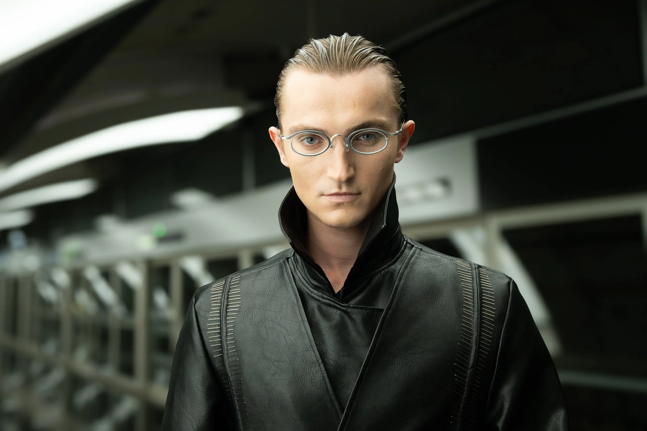 A young man with blond hair slicked back, wearing round silver glasses and a black leather jacket, looks directly at the camera in an urban setting with modern architecture.