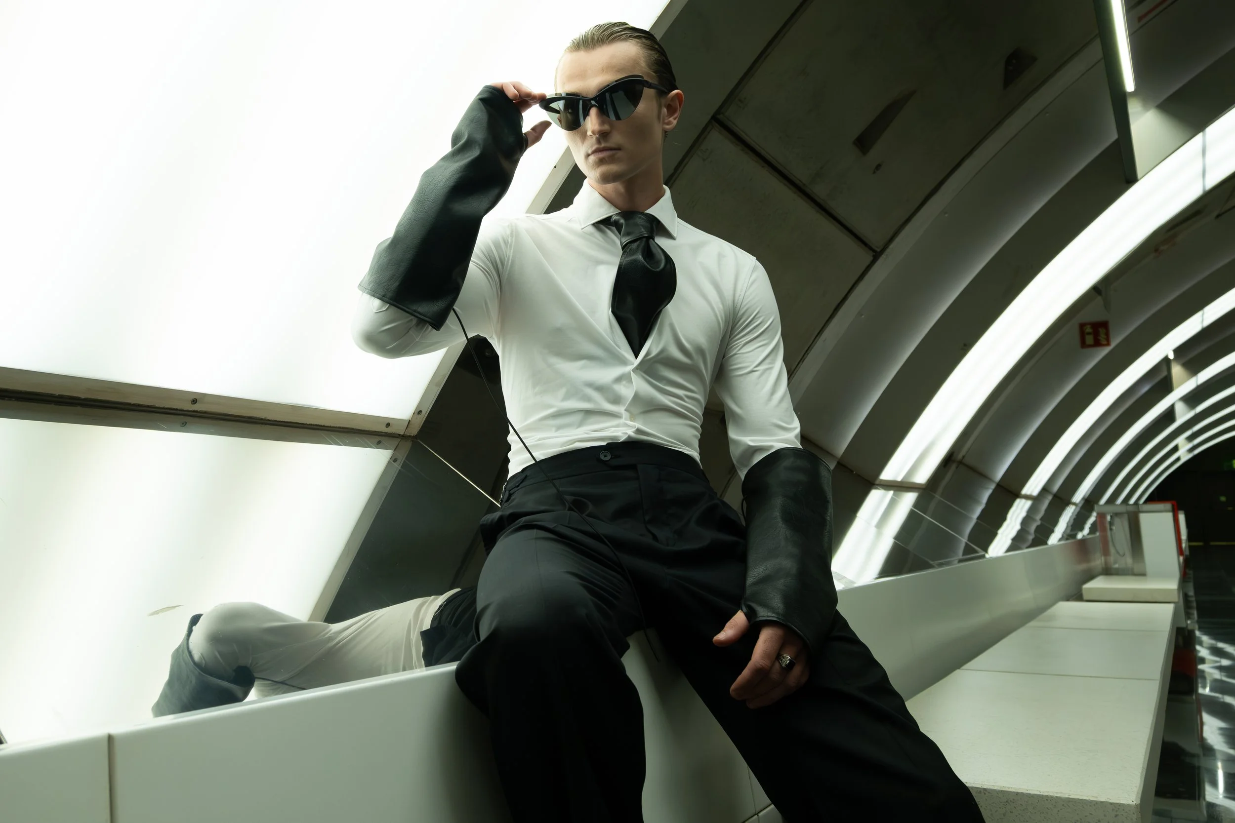 A man dressed in formal attire, wearing sunglasses and black leather gloves, sitting in an indoor space with curved architecture and bright lighting.