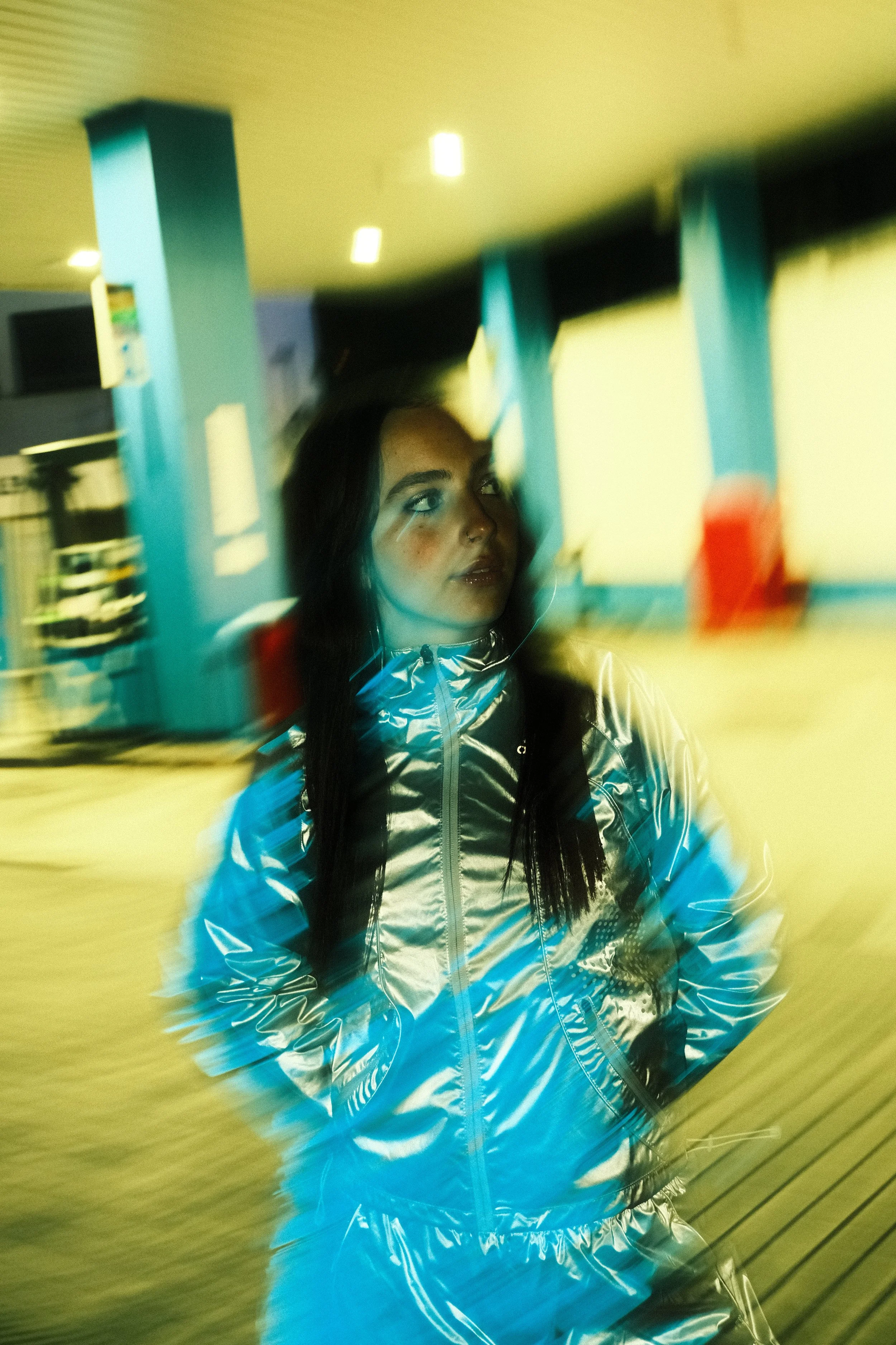 A young woman with long black hair wearing a shiny, metallic silver jacket stands in a futuristic indoor setting with teal columns and yellow walls. The image has a motion blur effect.