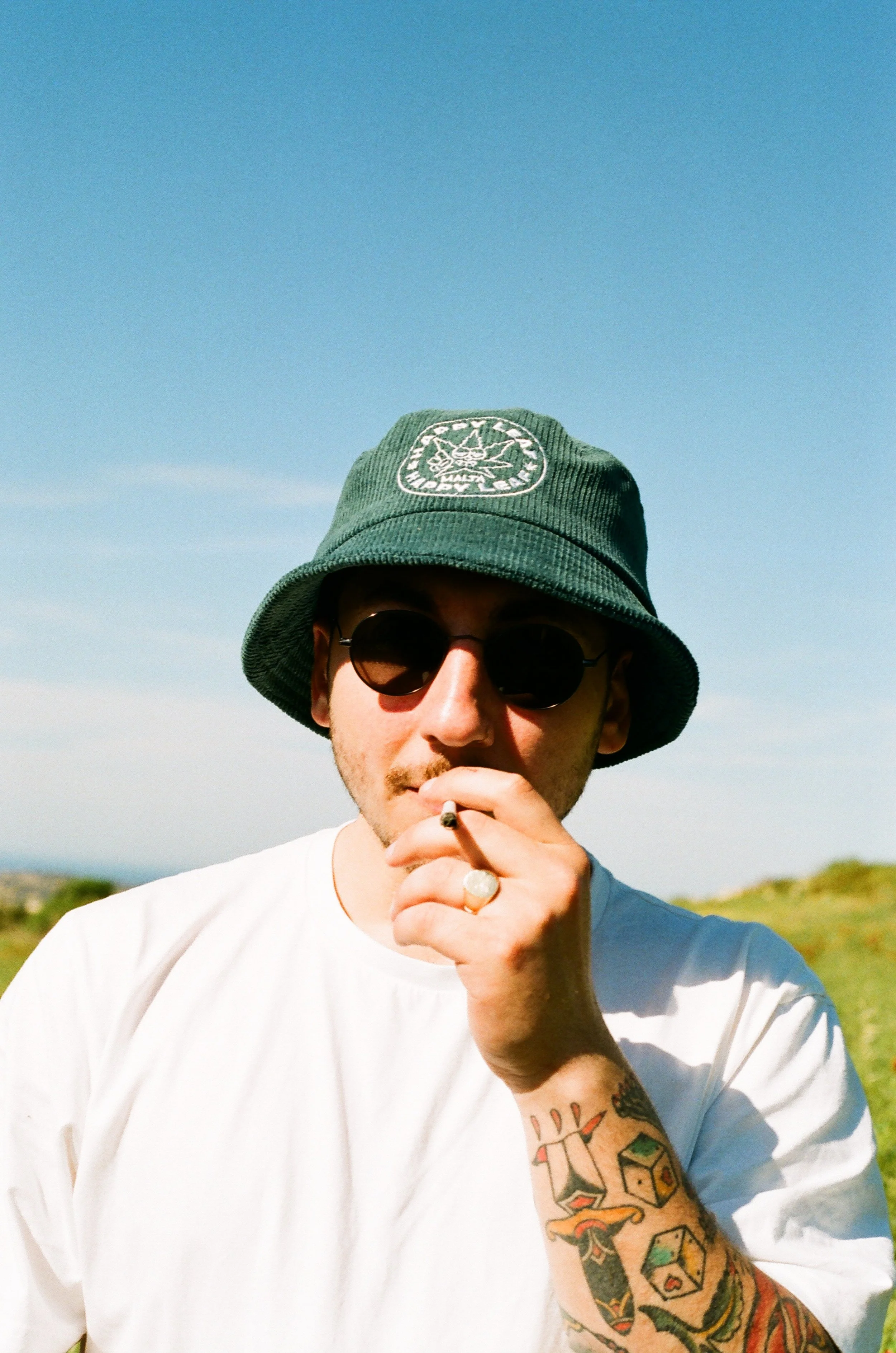A man wearing a green bucket hat with a patch, black sunglasses, a white T-shirt, and visible tattoos on his left arm, standing outdoors with a clear blue sky and greenery in the background, smoking a cigarette.
