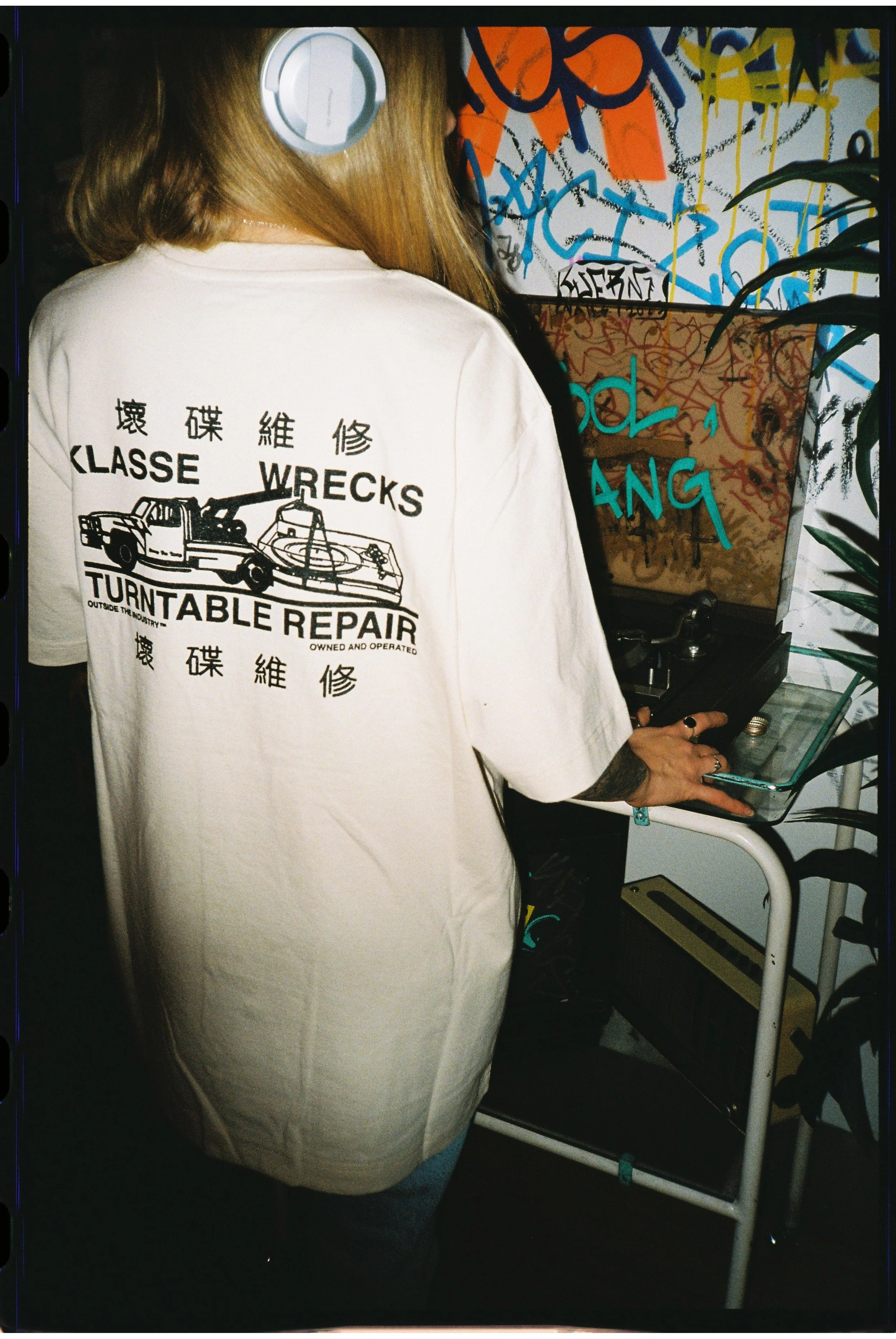 Person with long hair wearing a white T-shirt with black graphic design, including a wrecked car and turntable repair text, standing near a graffiti-covered wall and turntable.