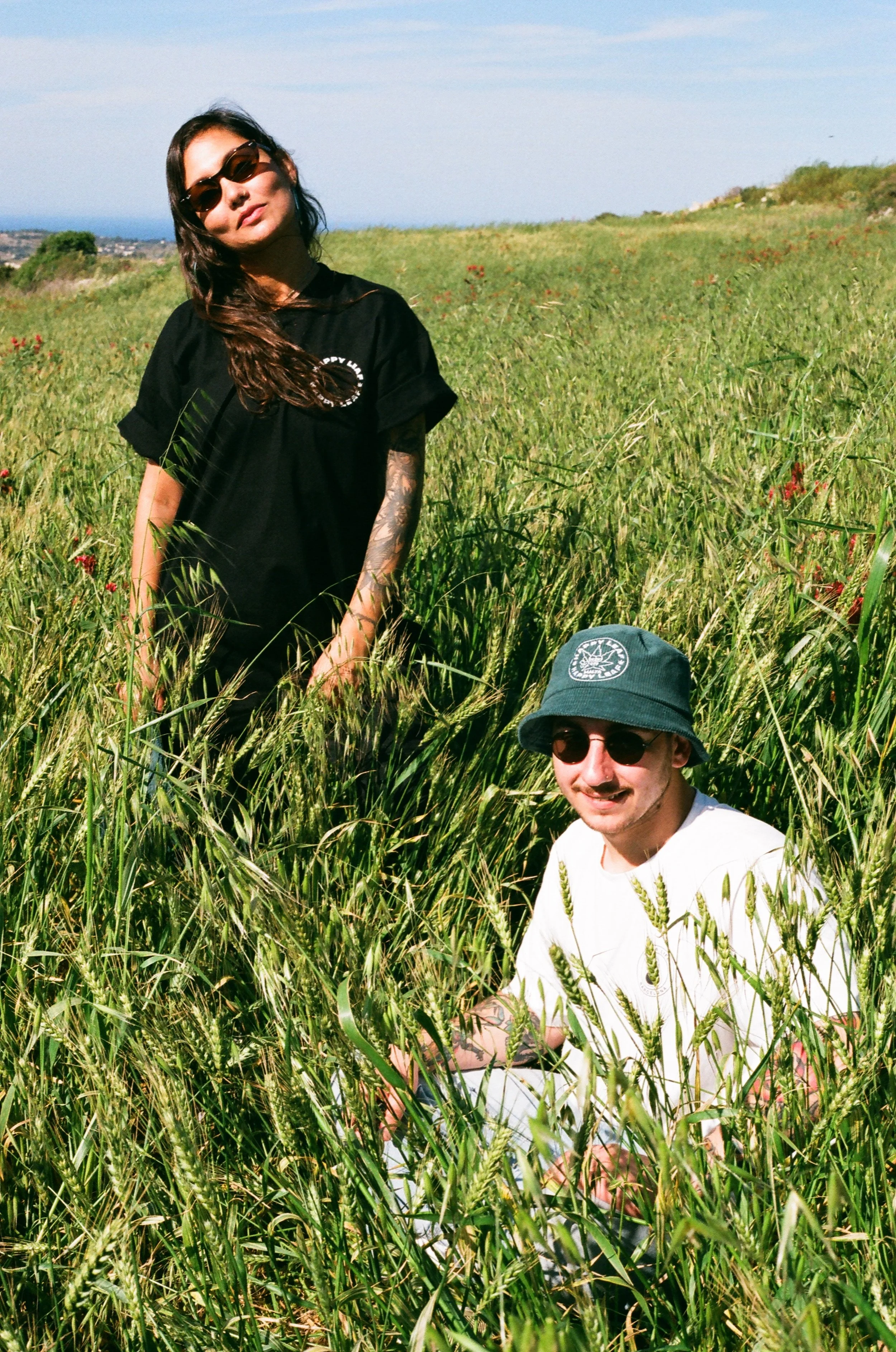 A woman with dark hair, wearing sunglasses and a black t-shirt, standing in a grassy field with a man wearing sunglasses and a bucket hat sitting in the grass beside her.