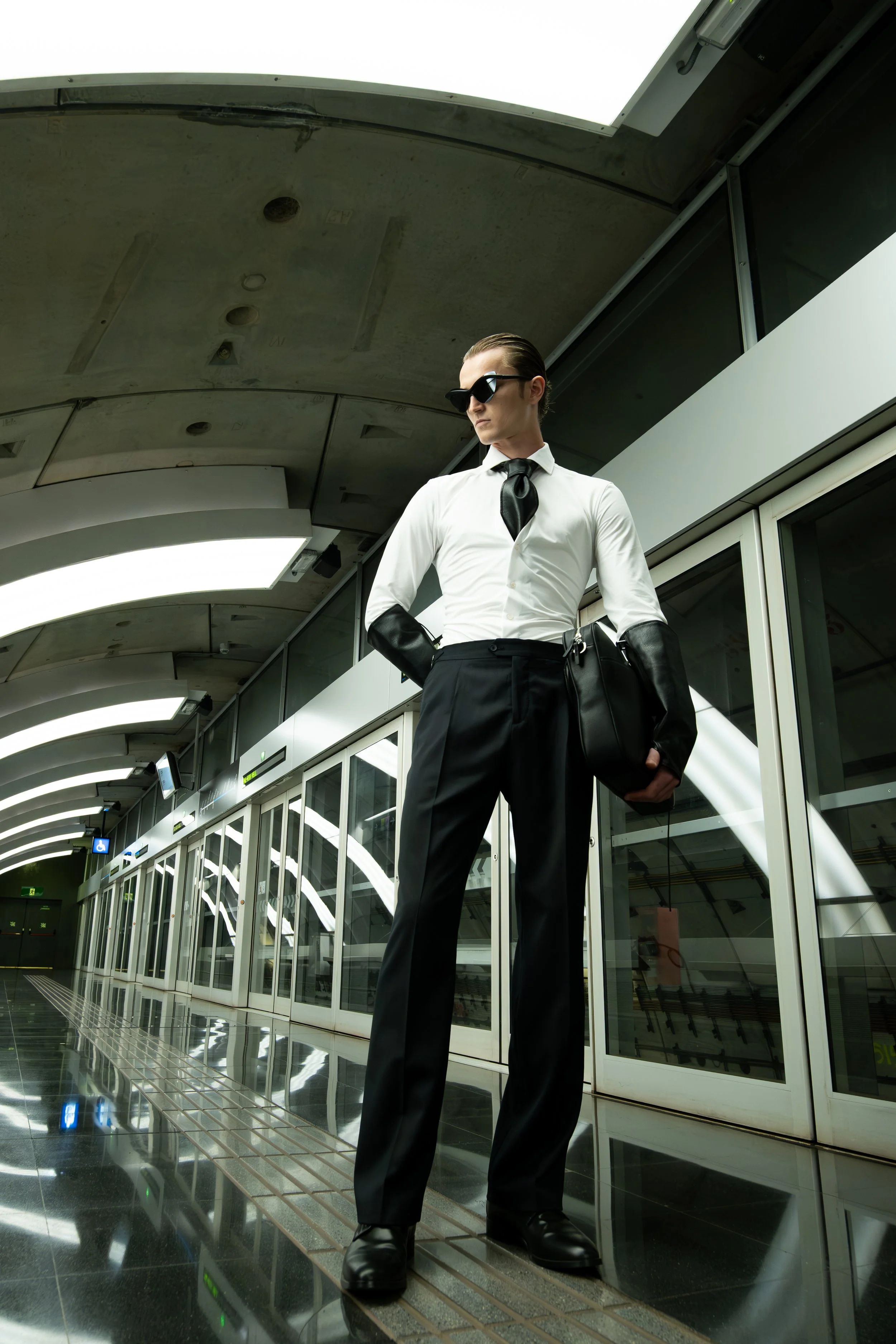 A person dressed in a white shirt, black tie, black gloves, and black sunglasses standing at an indoor modern airport or train station, looking down with a serious expression. The background features large glass windows and a curved ceiling with ligh