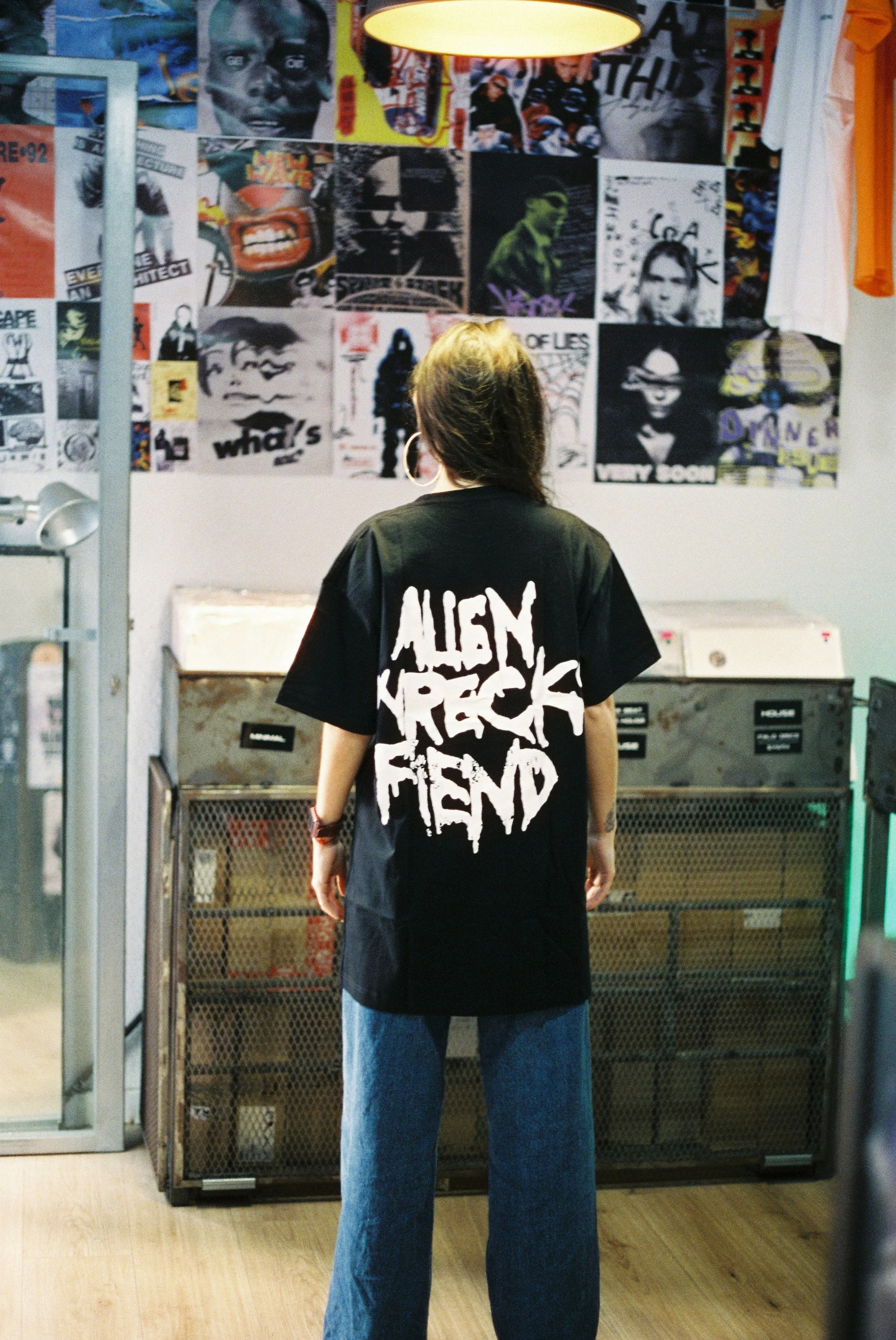 Person with long hair wearing a black t-shirt with white graffiti-style text, standing in front of vintage pop culture posters and arcade machines.