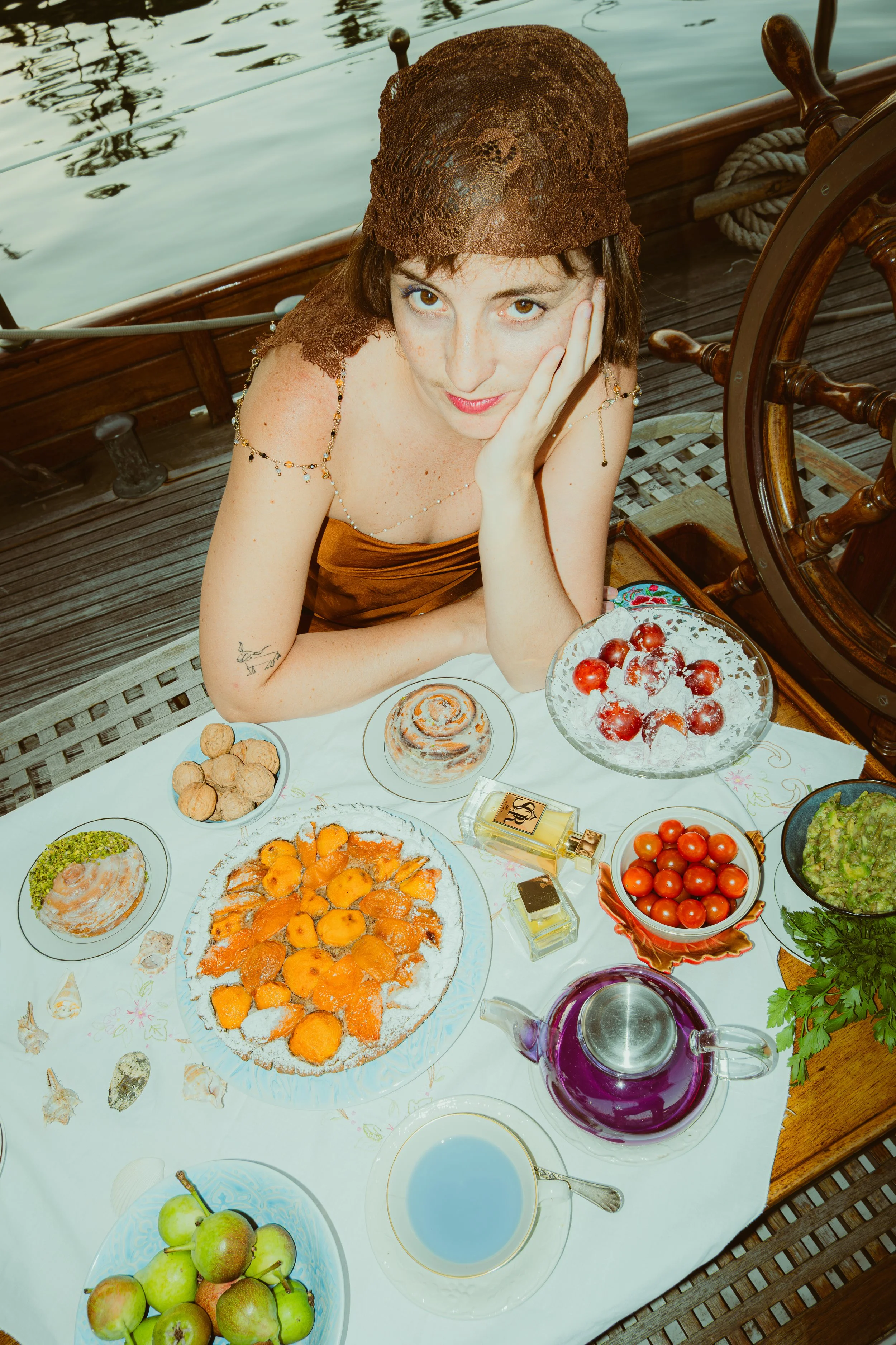 A woman with short dark hair and freckles, wearing a brown headscarf and dress, sitting at a table with various desserts and fruits on a boat near the water.
