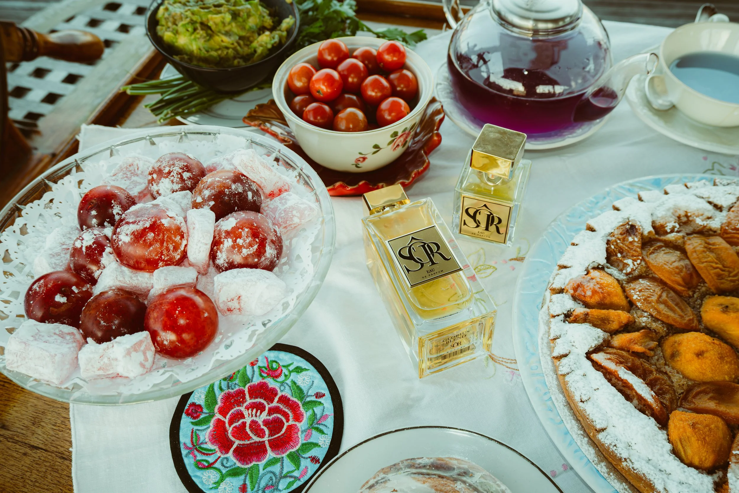 Decorative table with various dishes including a bowl of cherries, a plate of powdered-sugar-covered red fruits, cherry tomatoes, a pie dusted with powdered sugar, a jar of dark purple beverage, and perfume bottles.