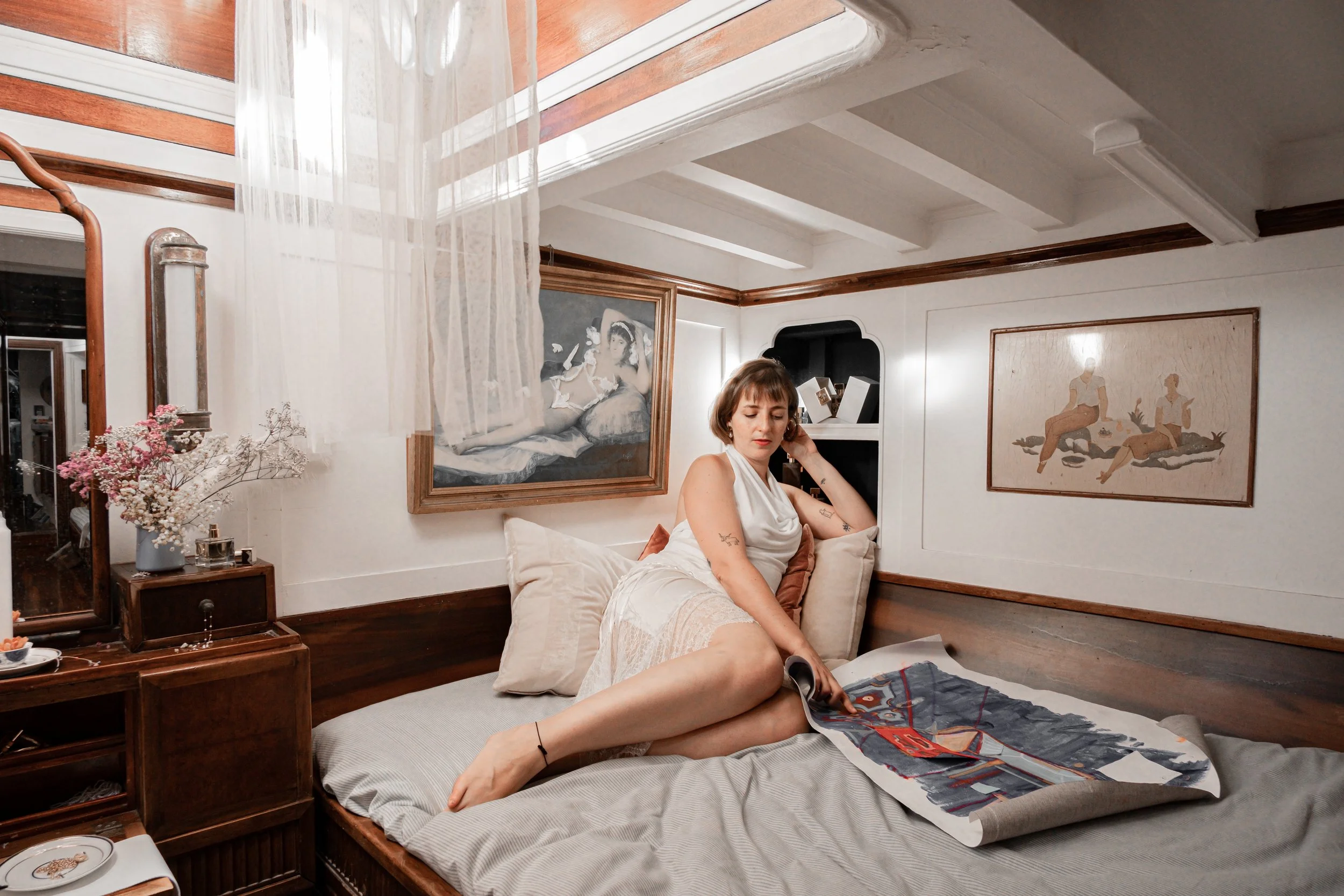 A woman with short brown hair and tattoos on her arms sits on a bed in a vintage-style bedroom, holding a phone to her ear. She is wearing a white lace top and beige shorts. The room features framed artwork on the walls, a wooden nightstand with pink
