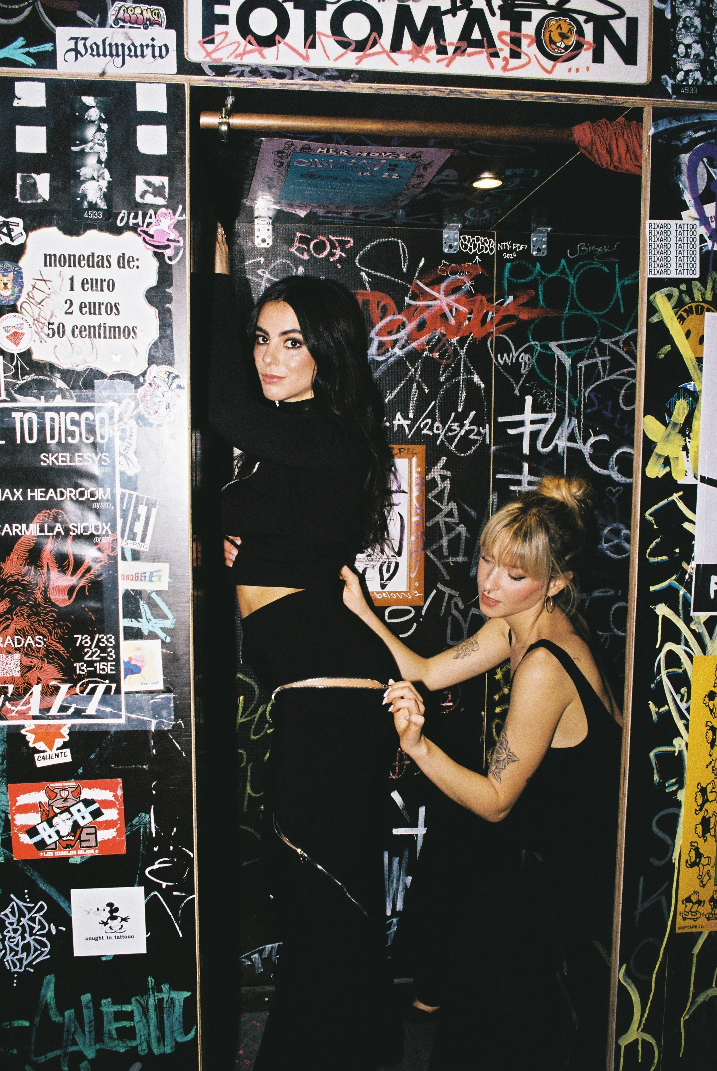 Two women in black outfits inside a graffiti-covered booth, one with dark hair and the other with blonde hair in a bun.