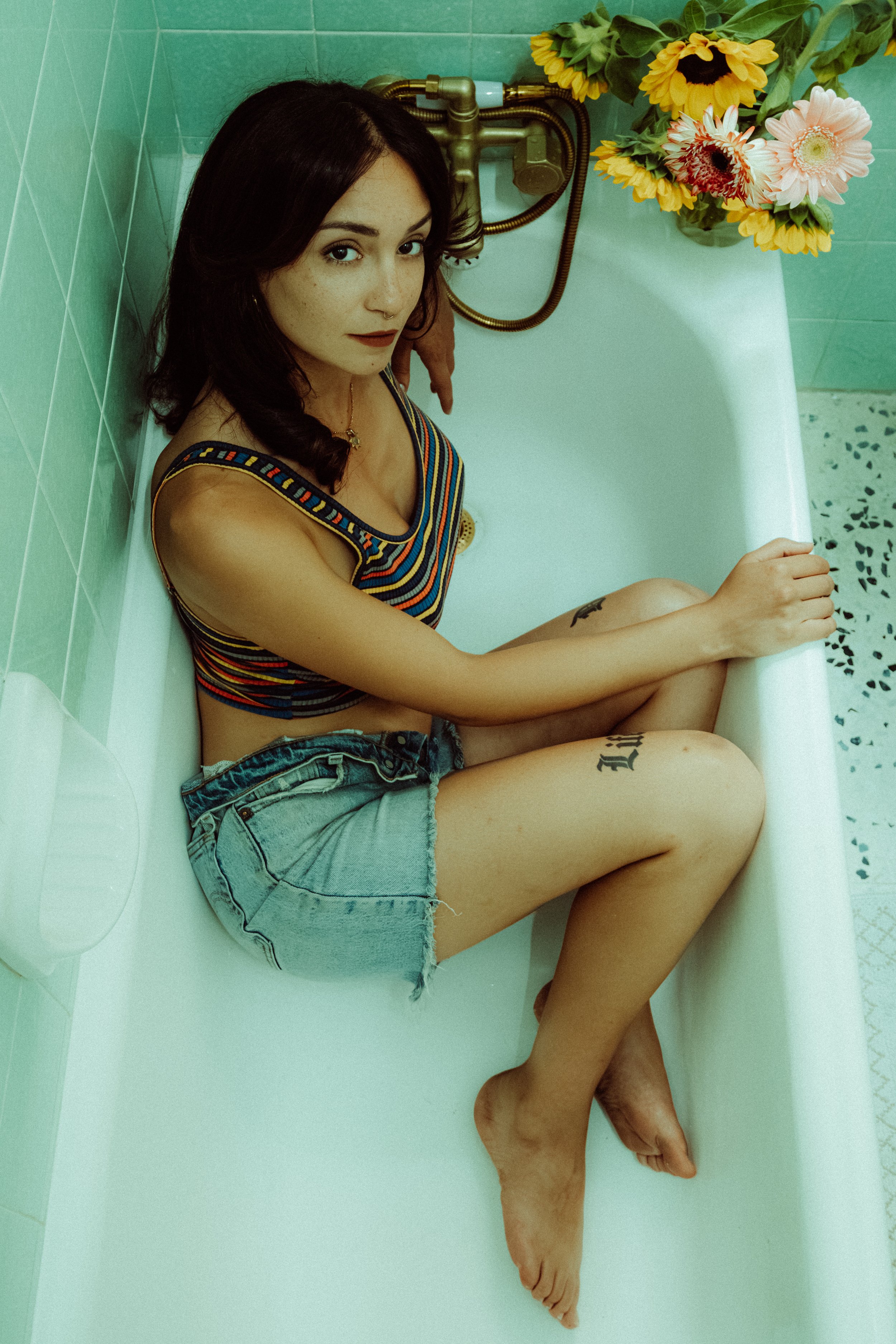A young woman with dark hair, freckles, and dark lipstick sitting in a vintage bathtub surrounded by green tiles and a bouquet of sunflowers and mixed flowers.