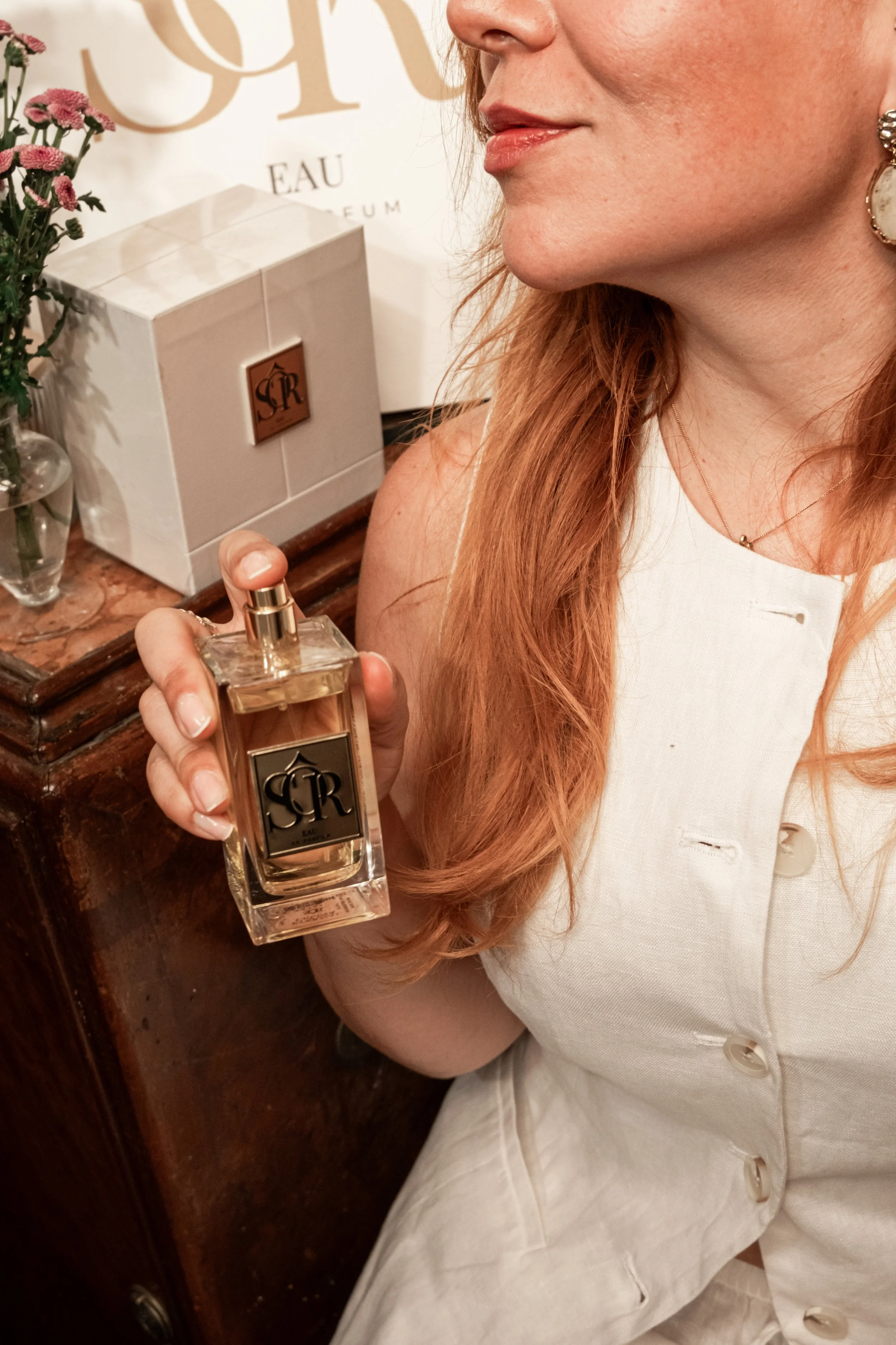 A woman with long, red hair holding a bottle of perfume near her shoulder, with a white box and pink flowers in the background.