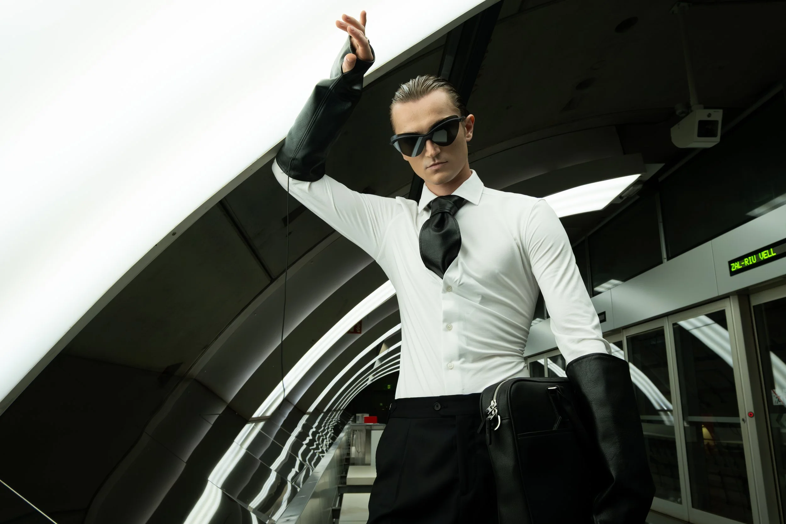 A man in a white dress shirt, black tie, and black pants standing in an airport terminal, adjusting his black sunglasses and holding a black bag.