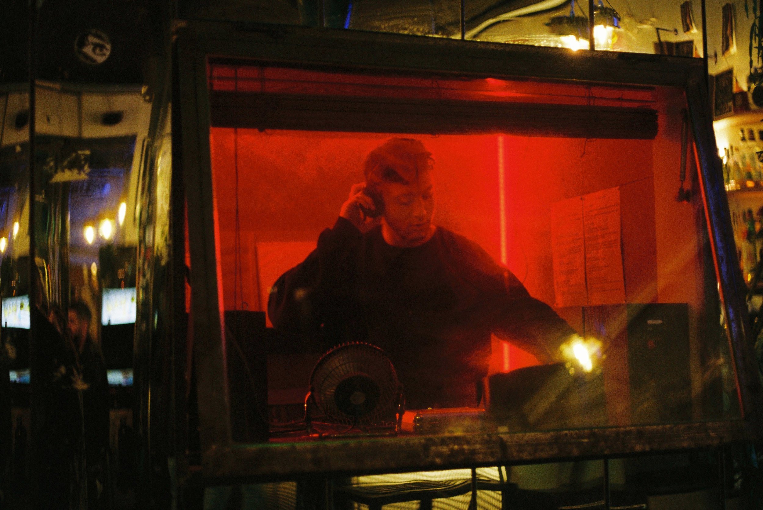 A DJ performs inside a glass booth illuminated with red lighting at a music venue.