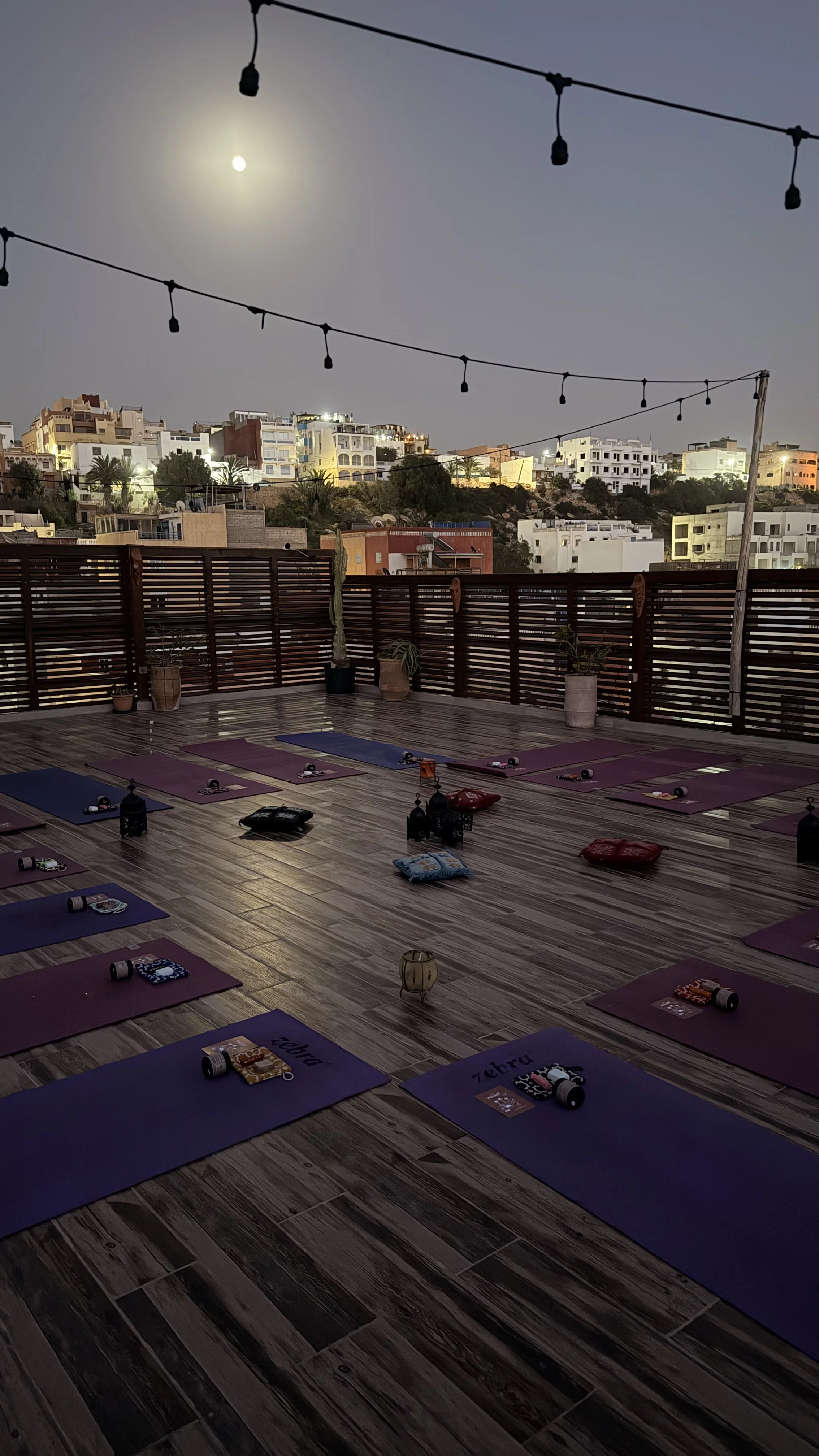 Une terrasse de yoga avec des tapis colorés, des coussins et des lanternes, surplombant une ville avec des bâtiments blancs éclairés par la lune, sous un ciel sombre avec des guirlandes lumineuses