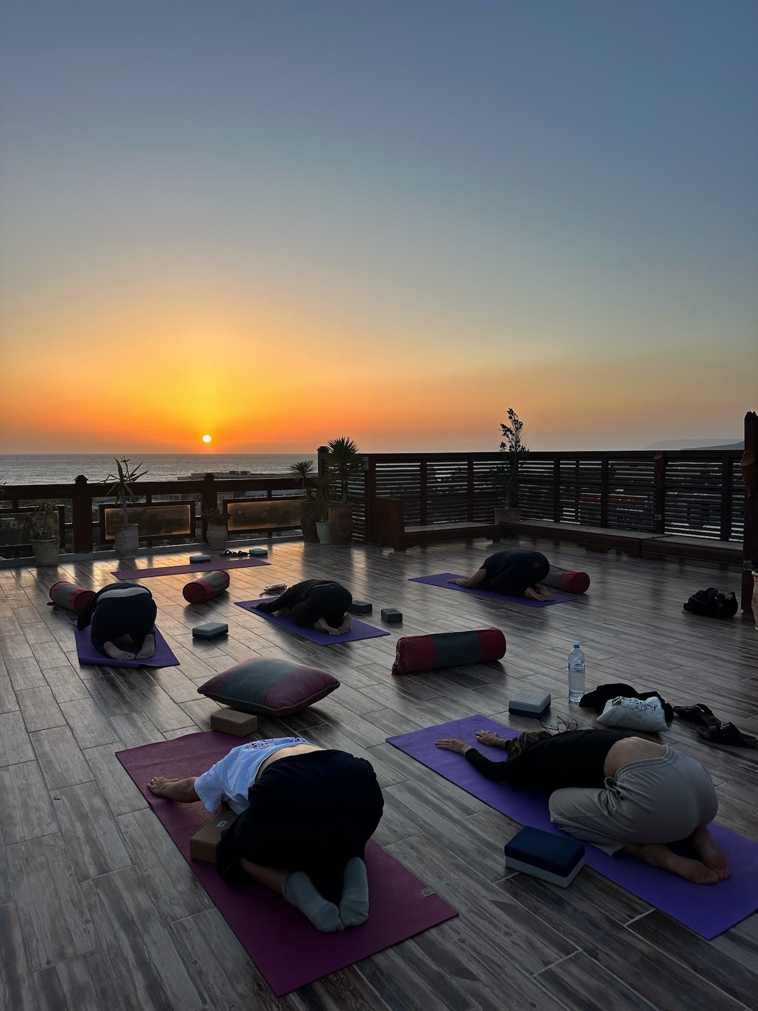 Groupe de personnes faisant du yoga sur un balcon au coucher du soleil, avec des matelas, des blocs de yoga et des coussins.
