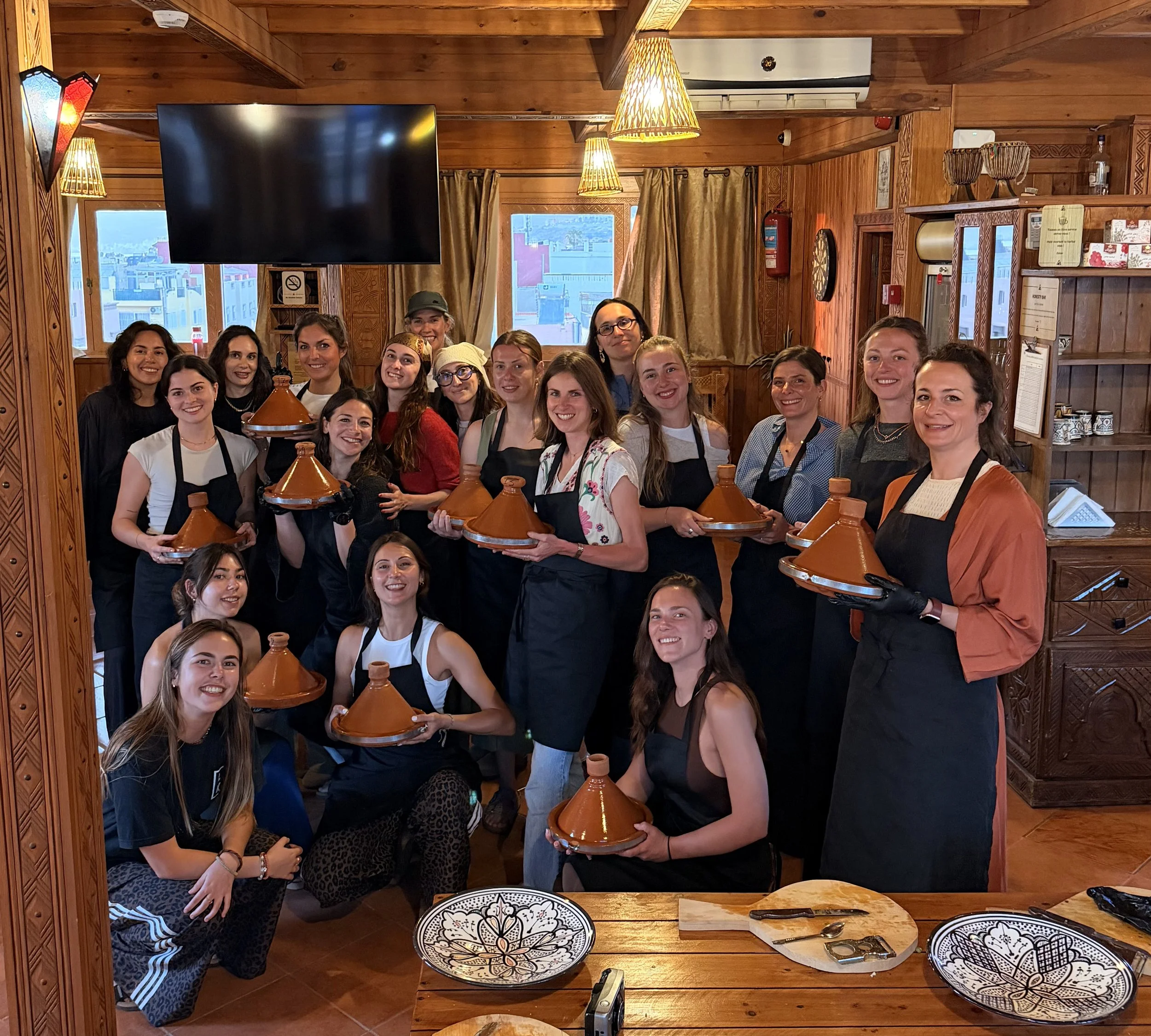 Groupe de femmes dans un restaurant en bois, tenant des tajines en terre cuite, organisant un atelier de cuisine marocaine.