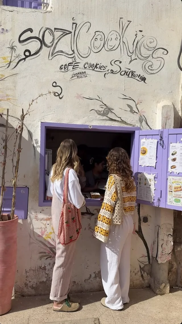 Deux femmes devant une petite fenêtre de café ou de boutique avec un mur peint en blanc et des affiches violettes, discutant ou commandant, avec des instructions ou menus affichés à côté.