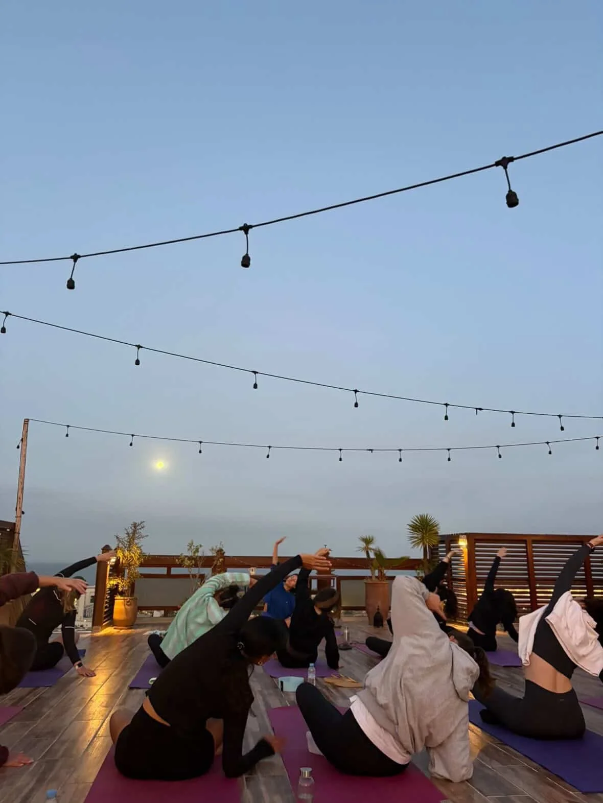 Groupe de personnes faisant du yoga sur un toit lors du coucher du soleil, avec des lampes suspendues au-dessus et la lune visible dans le ciel.