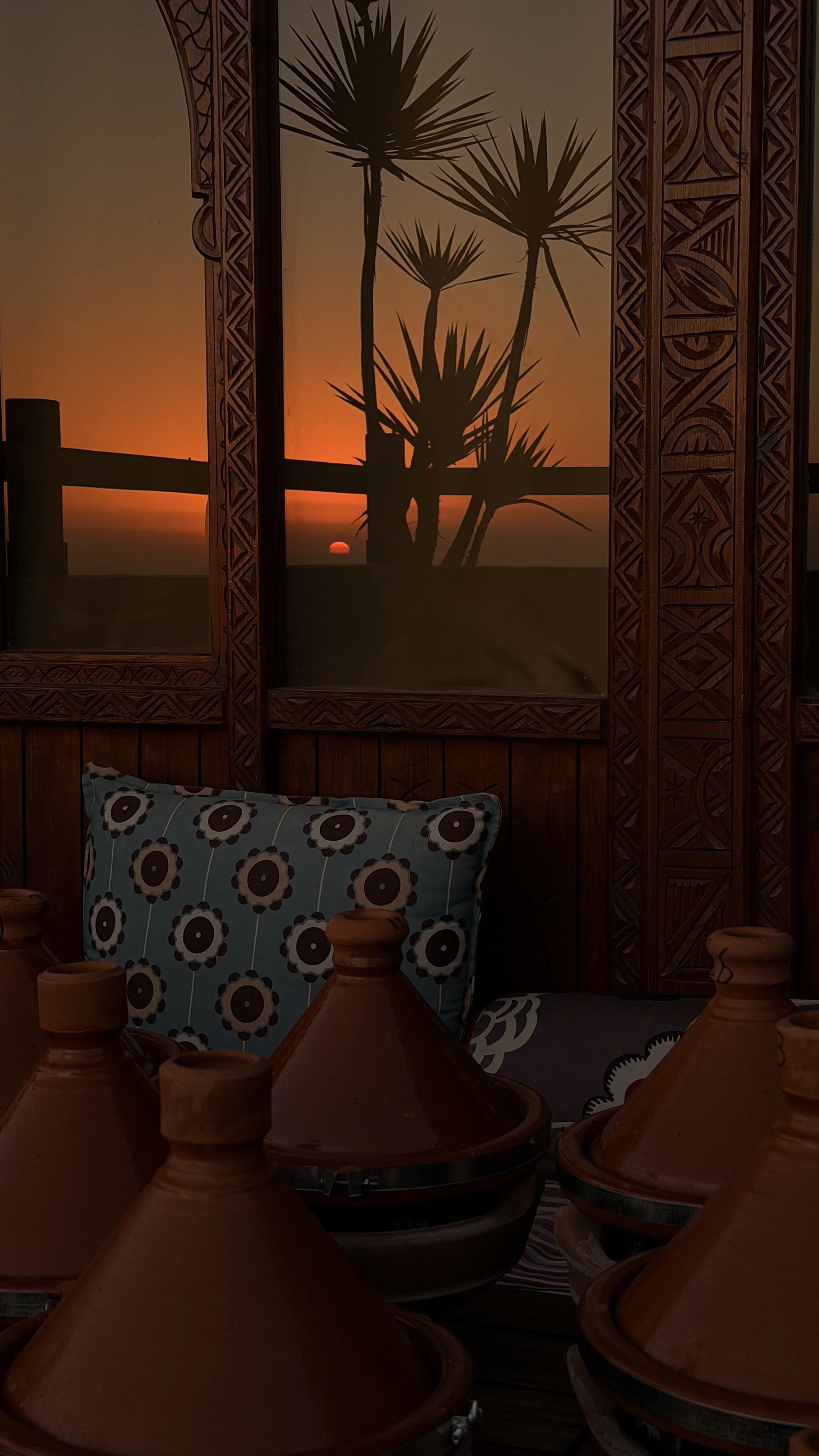 Coucher de soleil derrière une fenêtre en bois sculpté, avec une silhouette de palmier, dans un intérieur avec des coussins de style floral et des tajines en terre cuite.