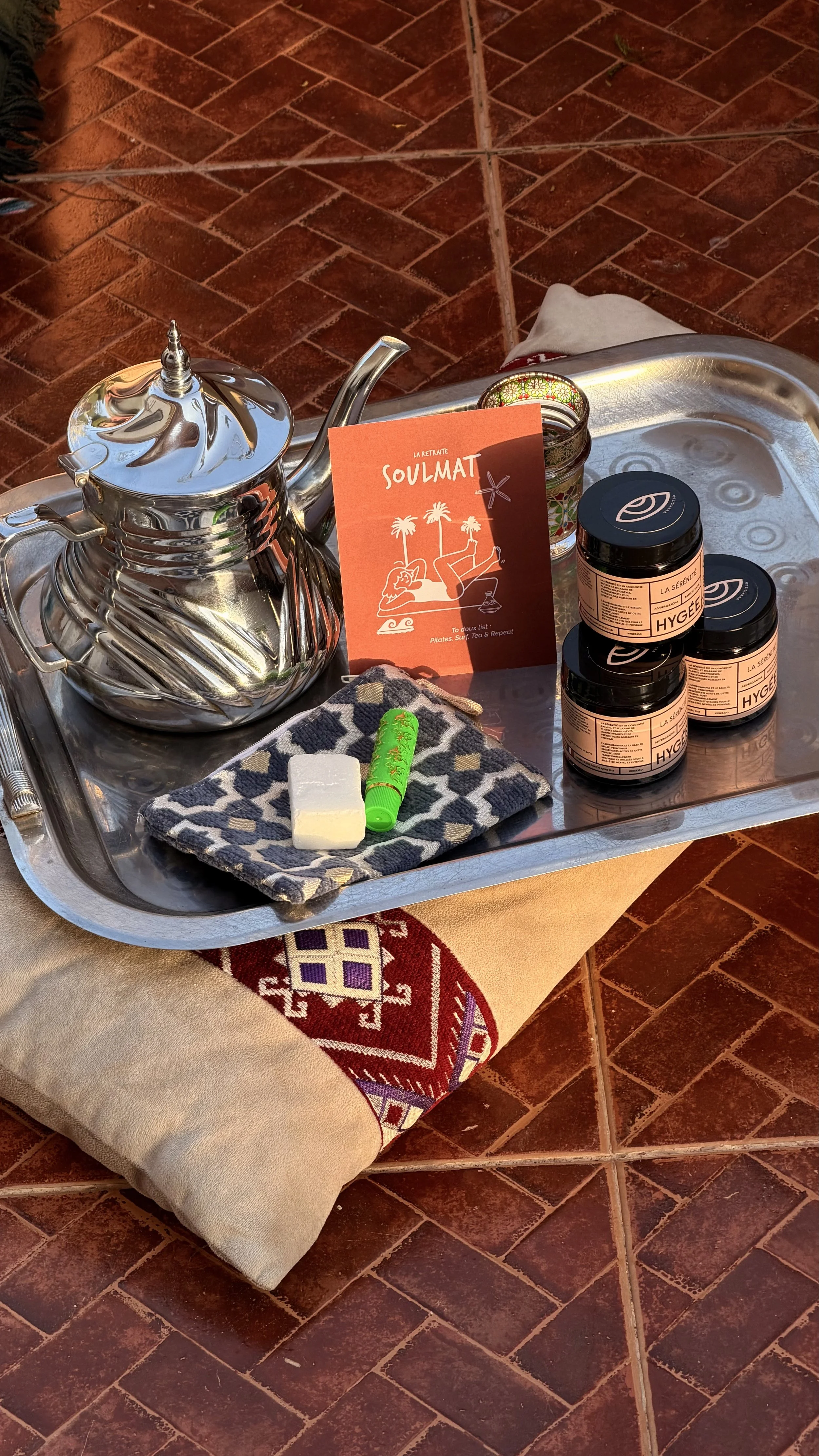 Un plateau en métal avec une théière en métal, un savon, un bâton de craie, une serviette avec un motif noir et blanc, trois petits pots de crème pour la peau, et une carte orange avec des palmiers et une femme allongée, sur un sol en carreaux de terre cuite.